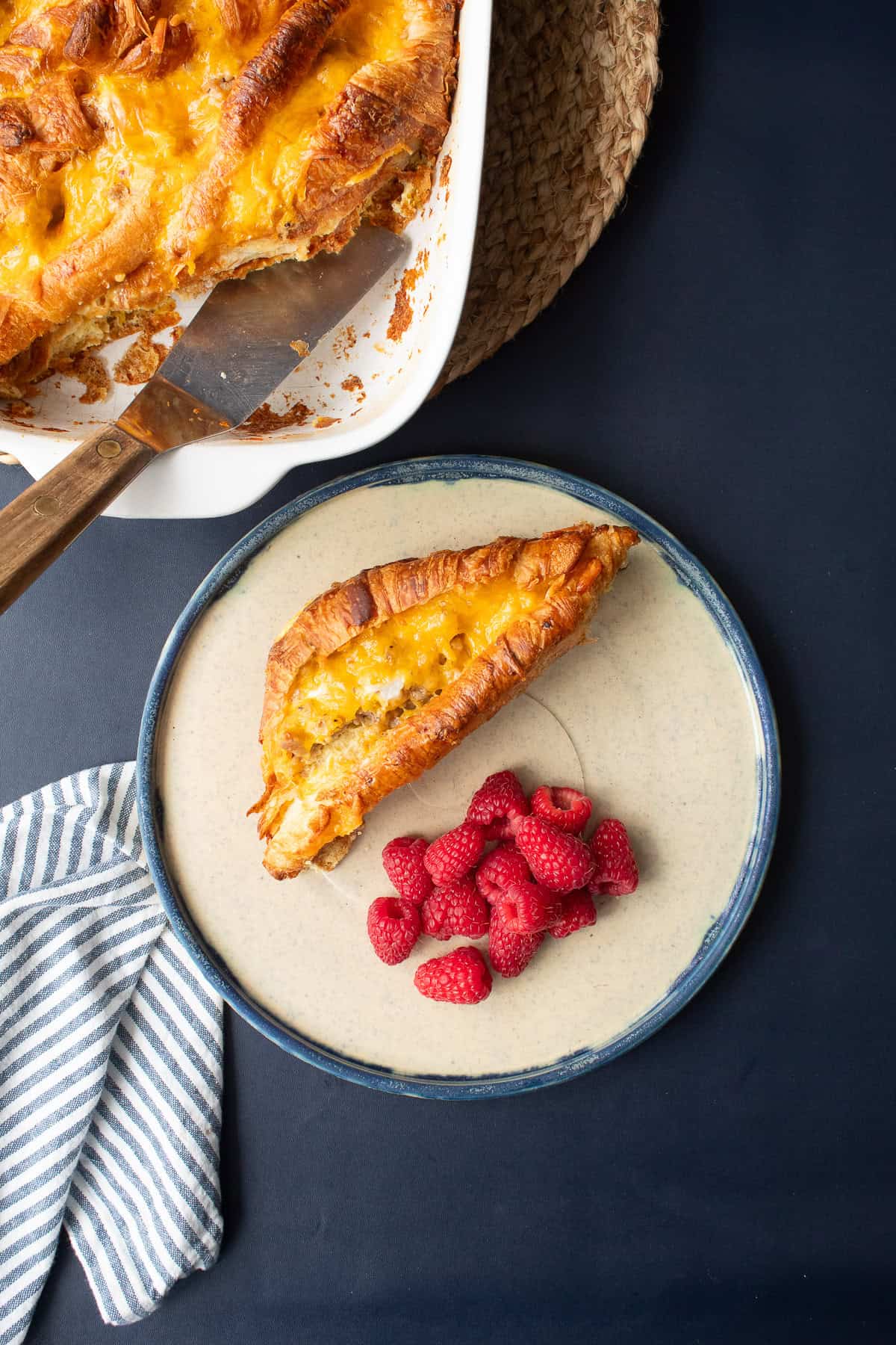 A baked croissant with sausage, egg, and cheese filling is served on a beige plate with a blue rim alongside some deep pink raspberries.