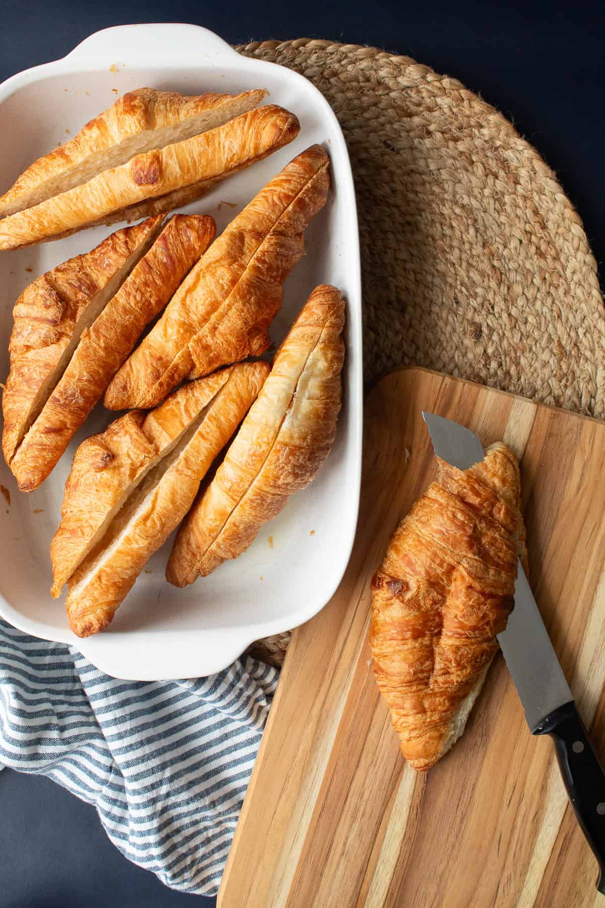 Croissants are sliced lengthwise with a serrated knife on a wooden cutting board.