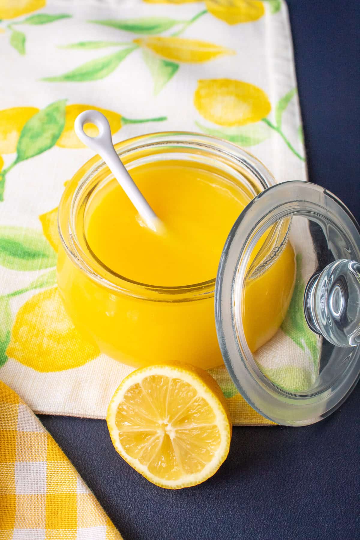 The bright yellow lemon curd is displayed in a glass jar with a white spoon.