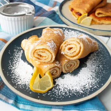 The rolled pancakes have a tender crepe-like texture and are dusted with powdered sugar on a blue plate garnished with a slice of lemon.
