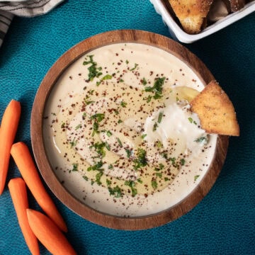 The lentil hummus is served in a wooden bowl and garnished with sumac, parsley, and a drizzle of olive oil alongside seasoned flatbread and carrots.