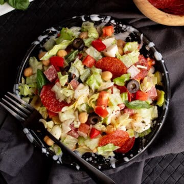 The mixed salad is plated on a black and white plate and has pieces of crispy pepperoni, chickpeas, olives, mozzarella, basil, pepper, and tomato visible.