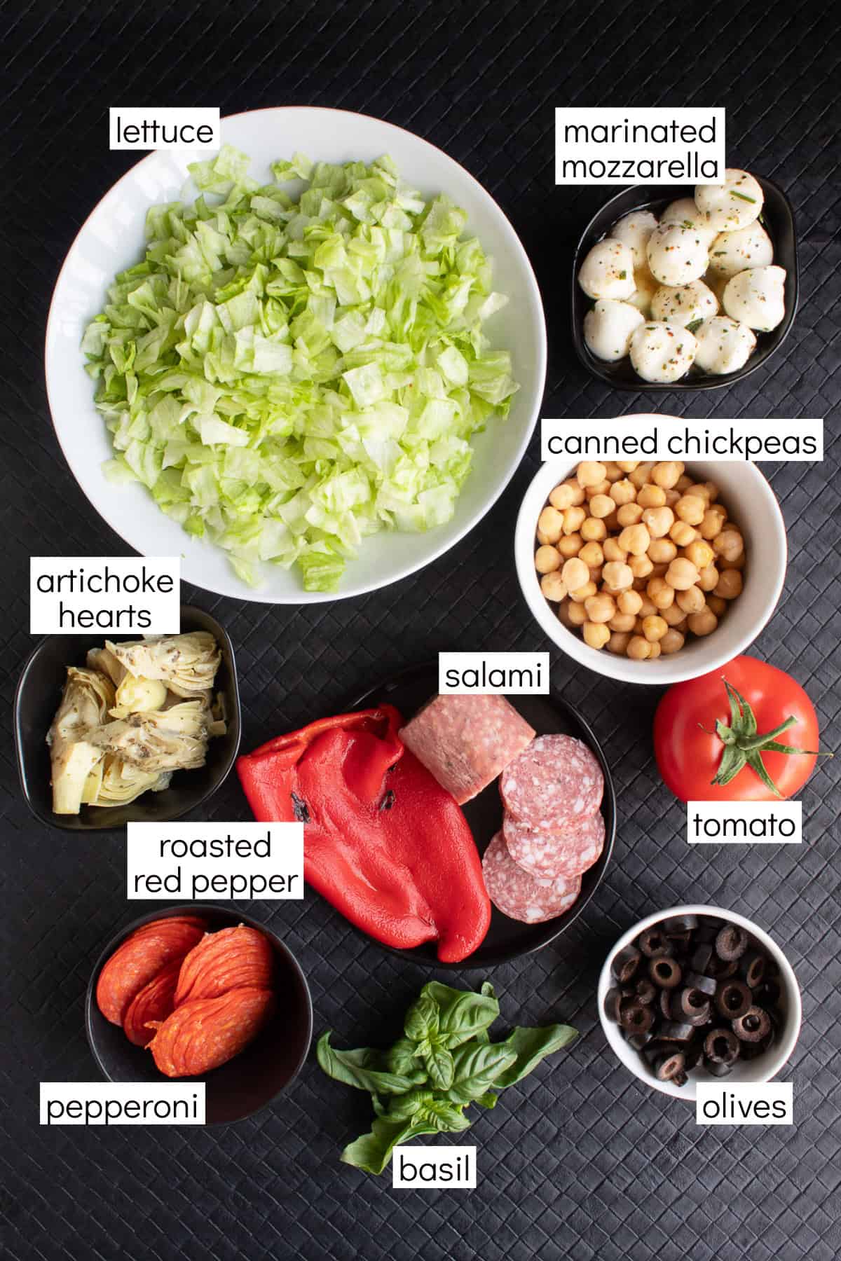 Ingredients for the recipe are displayed in various bowls on a black surface and are labeled with withe text boxes including lettuce, marinated mozzarella, canned chickpeas, salami, tomato, olives, basil, pepperoni, roasted red pepper, and artichoke hearts.