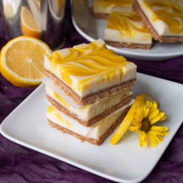 A stack of cheesecake bars sits on a white plate garnished with a slice of lemon and a small yellow flower.