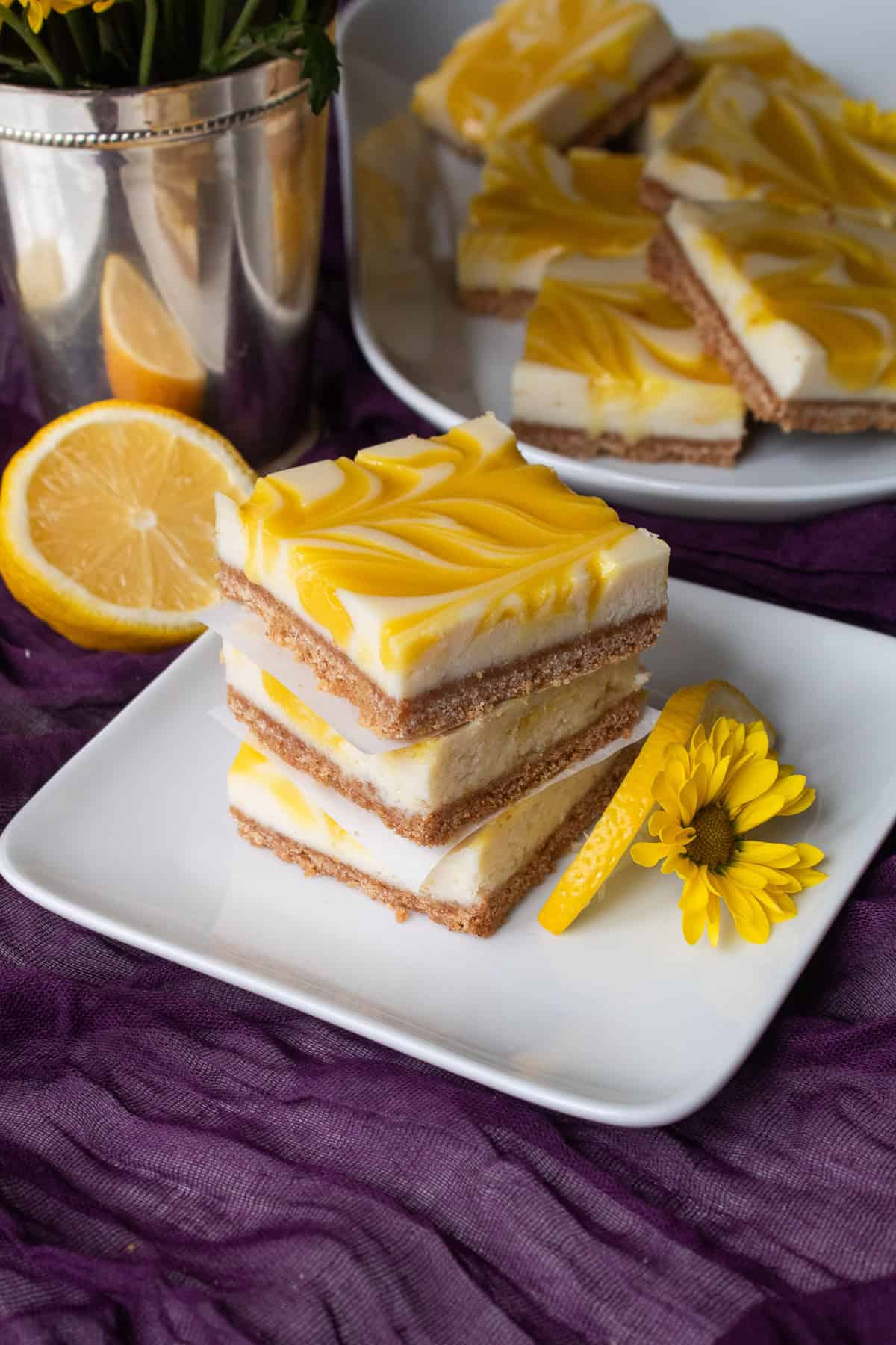 A stack of cheesecake bars sits on a white plate garnished with a slice of lemon and a small yellow flower.
