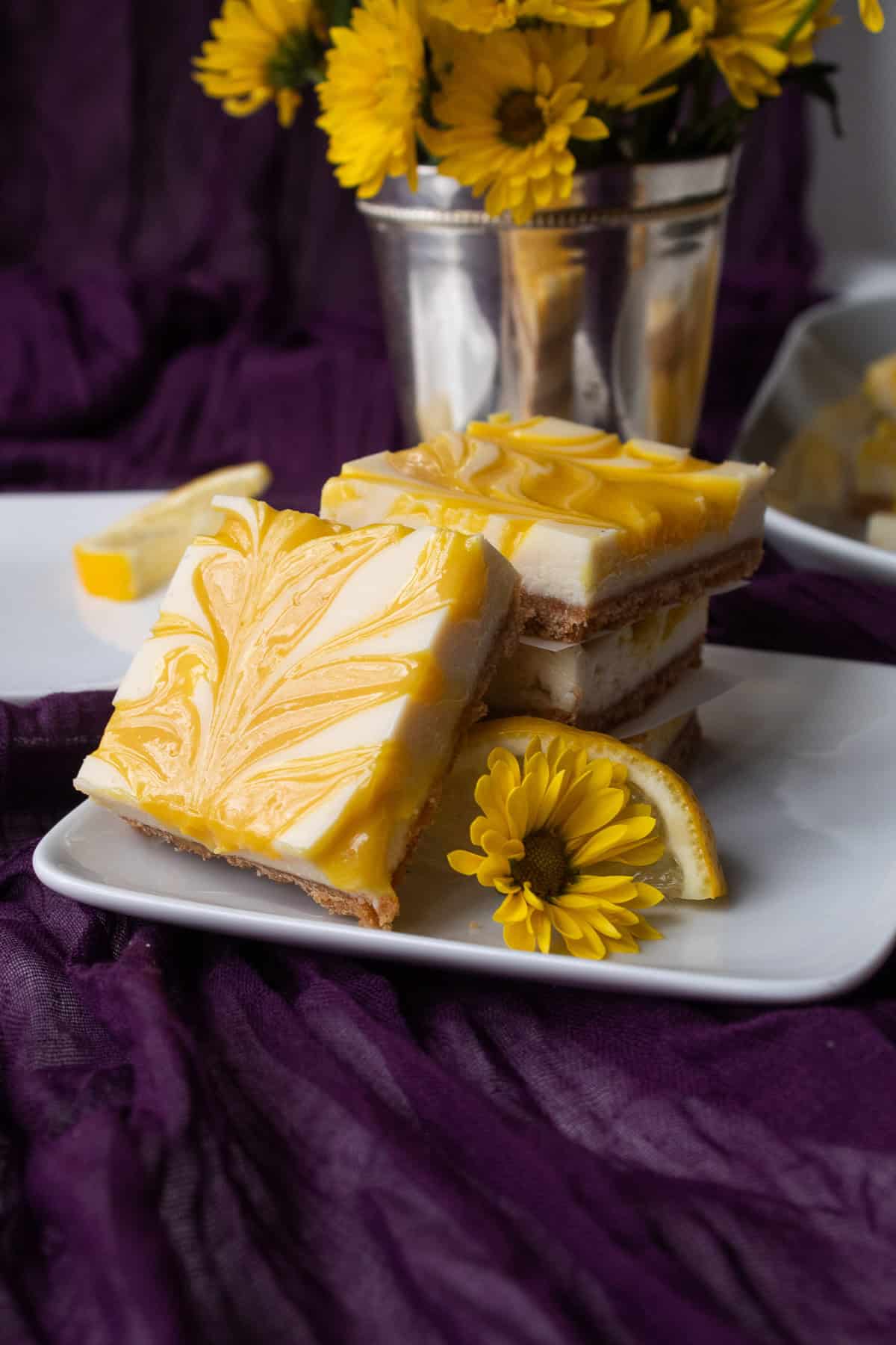 A plate of cheesecake bars with marbled yellow lemon curd sits on a purple background and is garnished with a small yellow flower and a lemon slice.