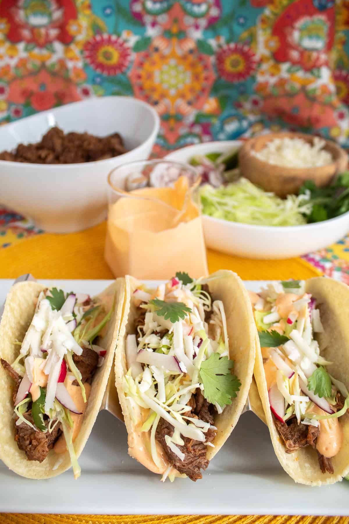 Three tacos filled with shredded beef chuck, chipotle crema, and fresh toppings are shown on a white platter.