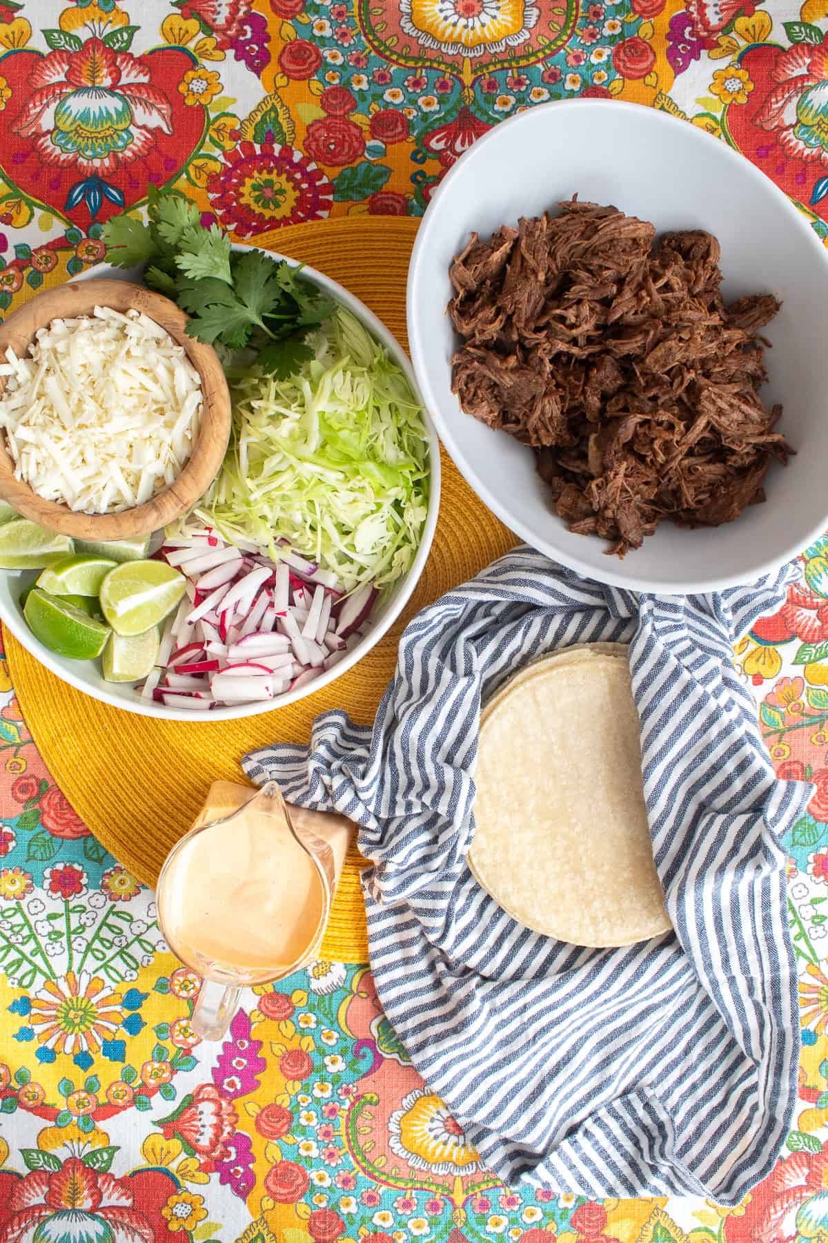 Components for the tacos, including shredded beef, steamed tortillas, chipotle crema, cabbage, radishes, cheese, limes, and cilantro, are displayed in bowls.