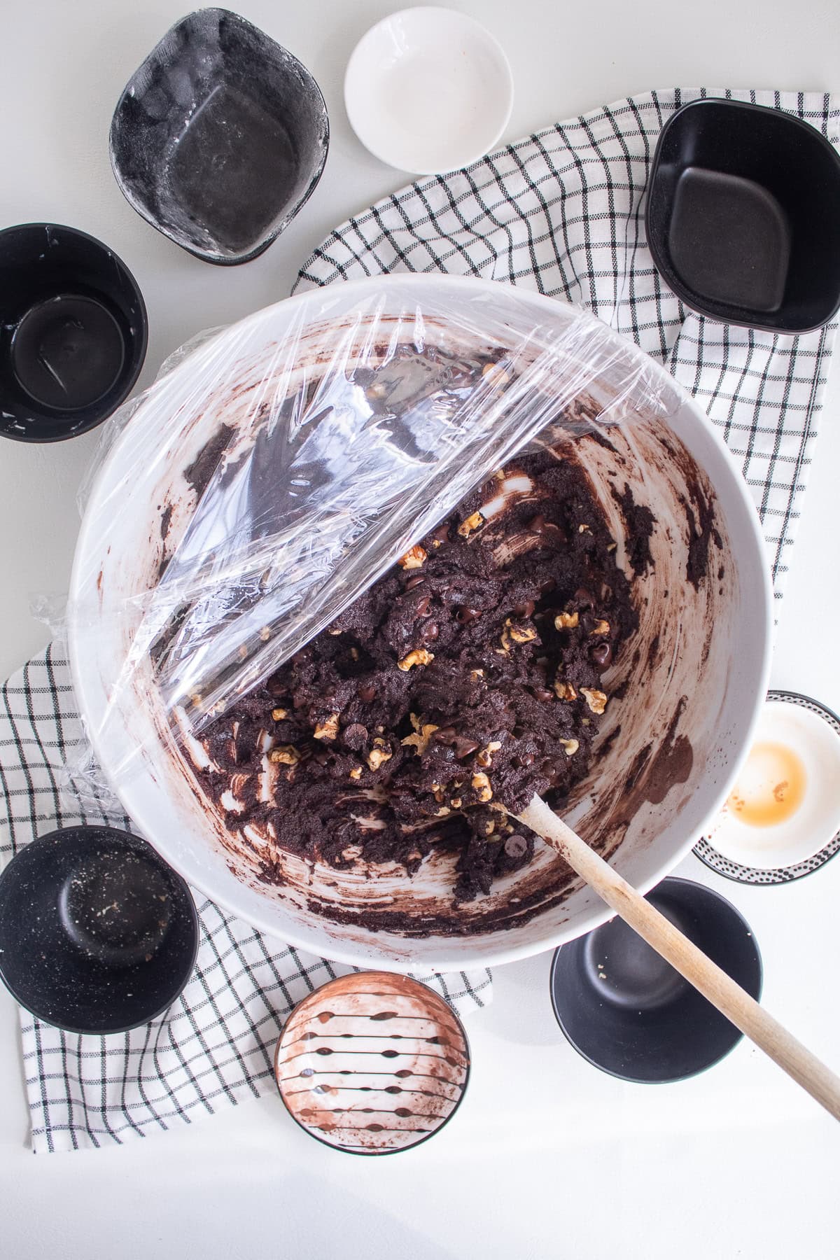 The white mixing bowl containing the finished cookie dough is covered with plastic wrap.