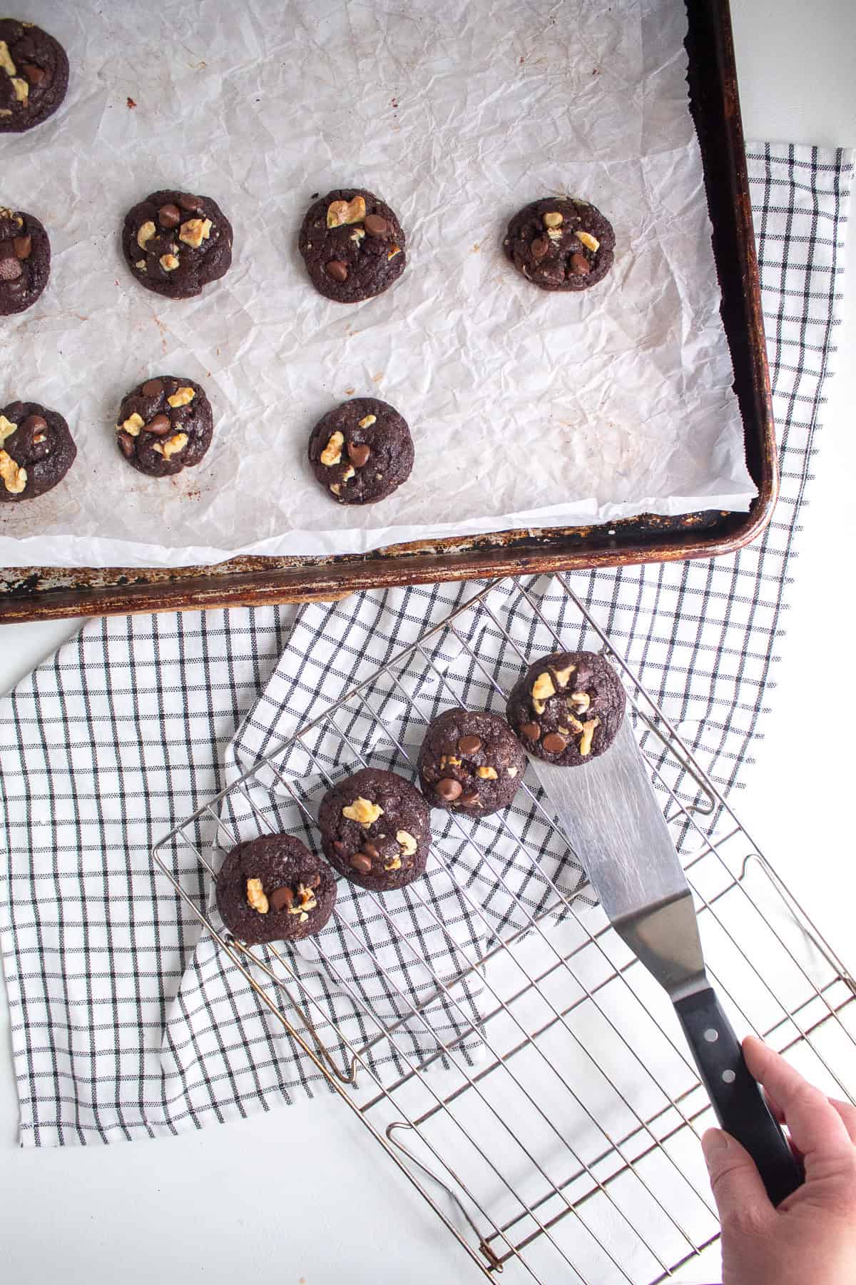 Cookies are being transferred from a parchment paper lined sheet pan to a wire cooling rack with a metal spatula.