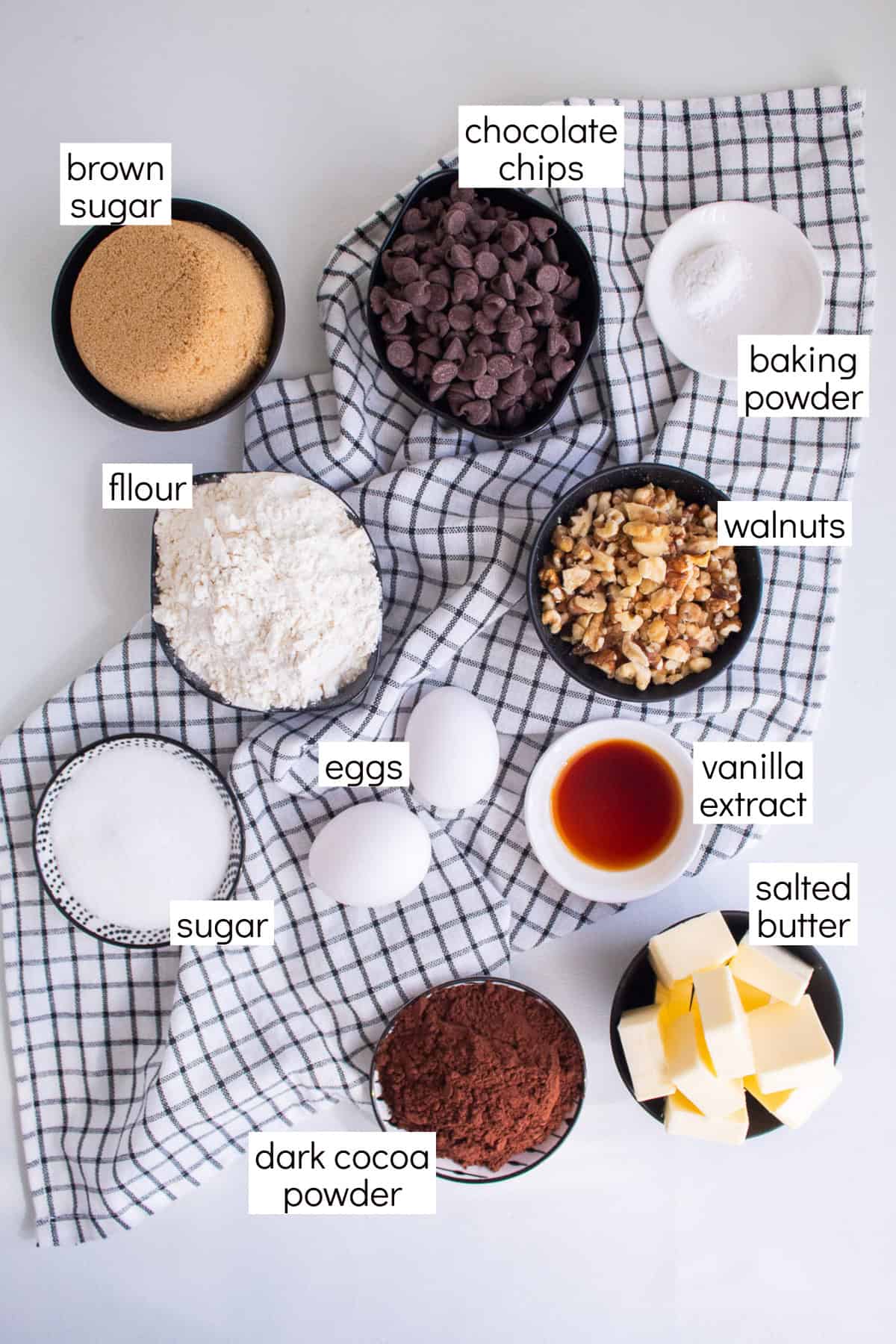 Ingredients for the recipe are displayed on a black and white cloth and are labeled with white text boxes including brown sugar, chocolate chips, baking powder, walnuts, vanilla extract, salted butter, dark cocoa powder, sugar, eggs, and flour.