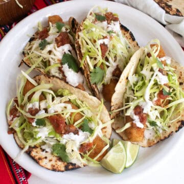 A platter of four fish stick tacos are garnished with white sauce, cabbage, salsa, and cilantro.