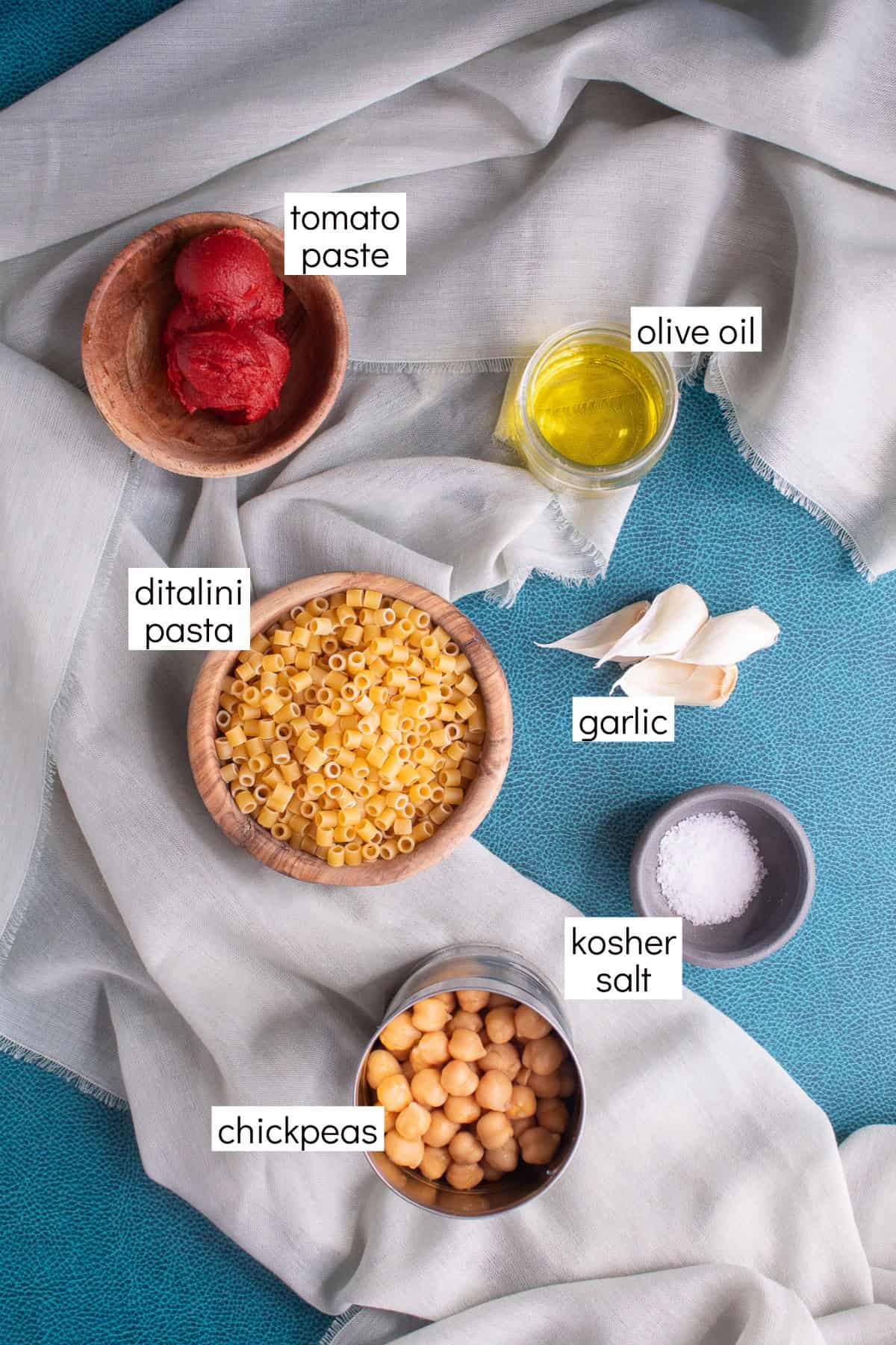 Ingredients for the recipe are displayed on a gray and blue background and have white text box labels including tomato paste, olive oil, ditalini pasta, garlic, kosher salt, and chickpeas.