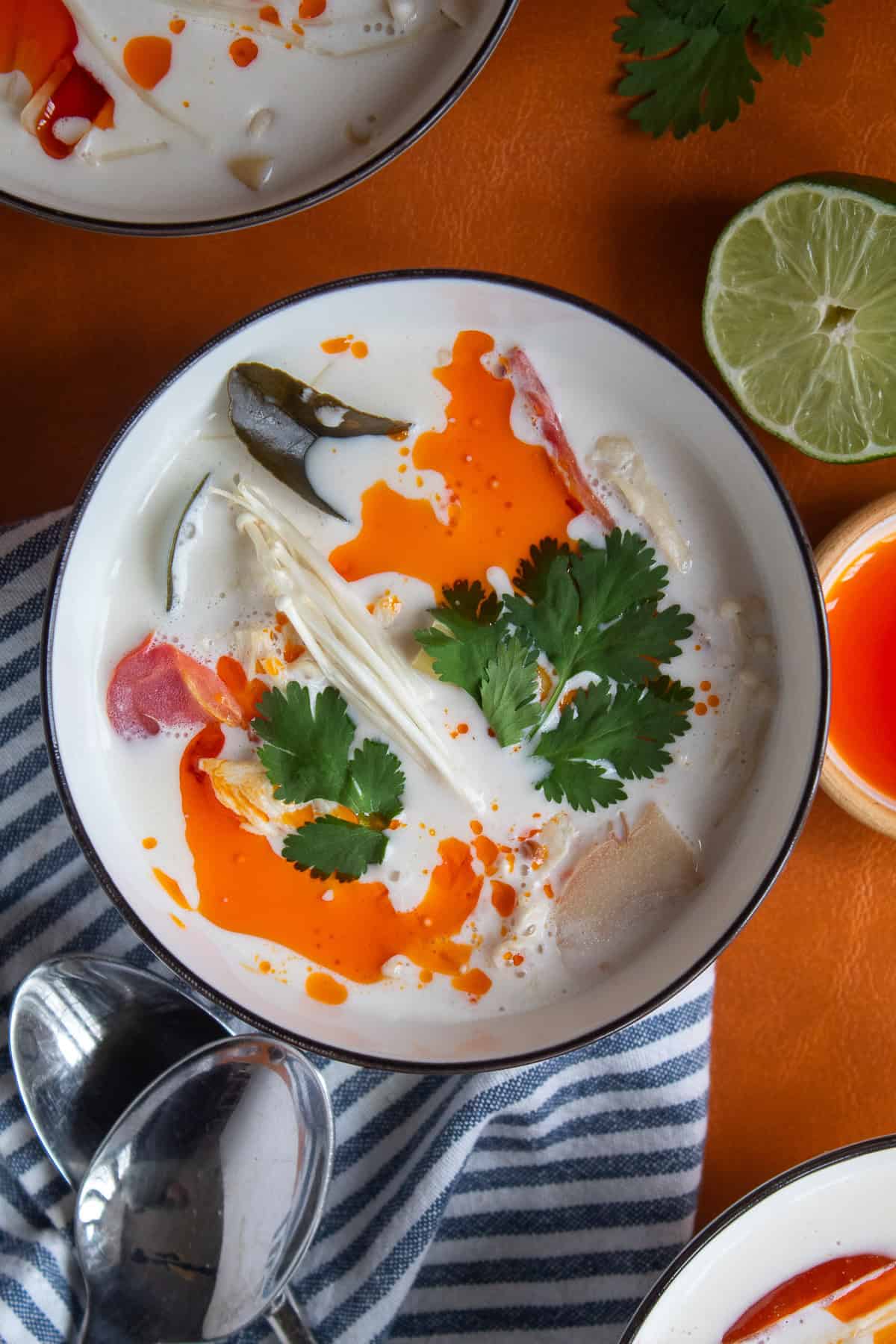 A bowl of white soup garnished with chili oil, cilantro, and mushrooms sits on blue striped cloth on an orange background.