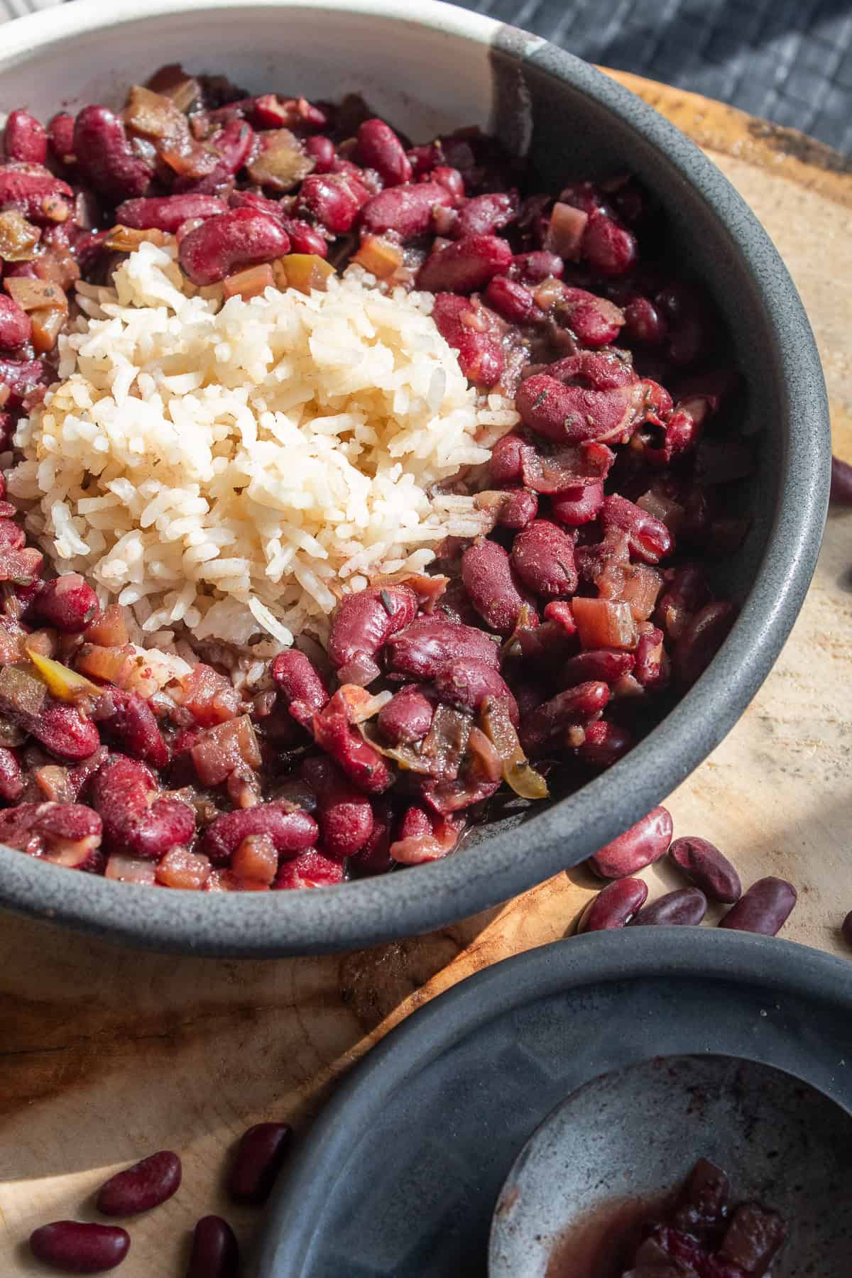 A bowl of red beans and rice is bathed in sunlight and sits on a wooden background alongside a spoon of cooked beans.