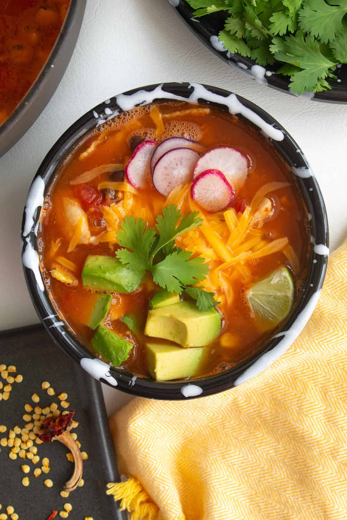 A bowl of garnished brothy soup sits on a white surface alongside the pot of soup, a plate of toppings, and the seeds and stem from a dried chile.