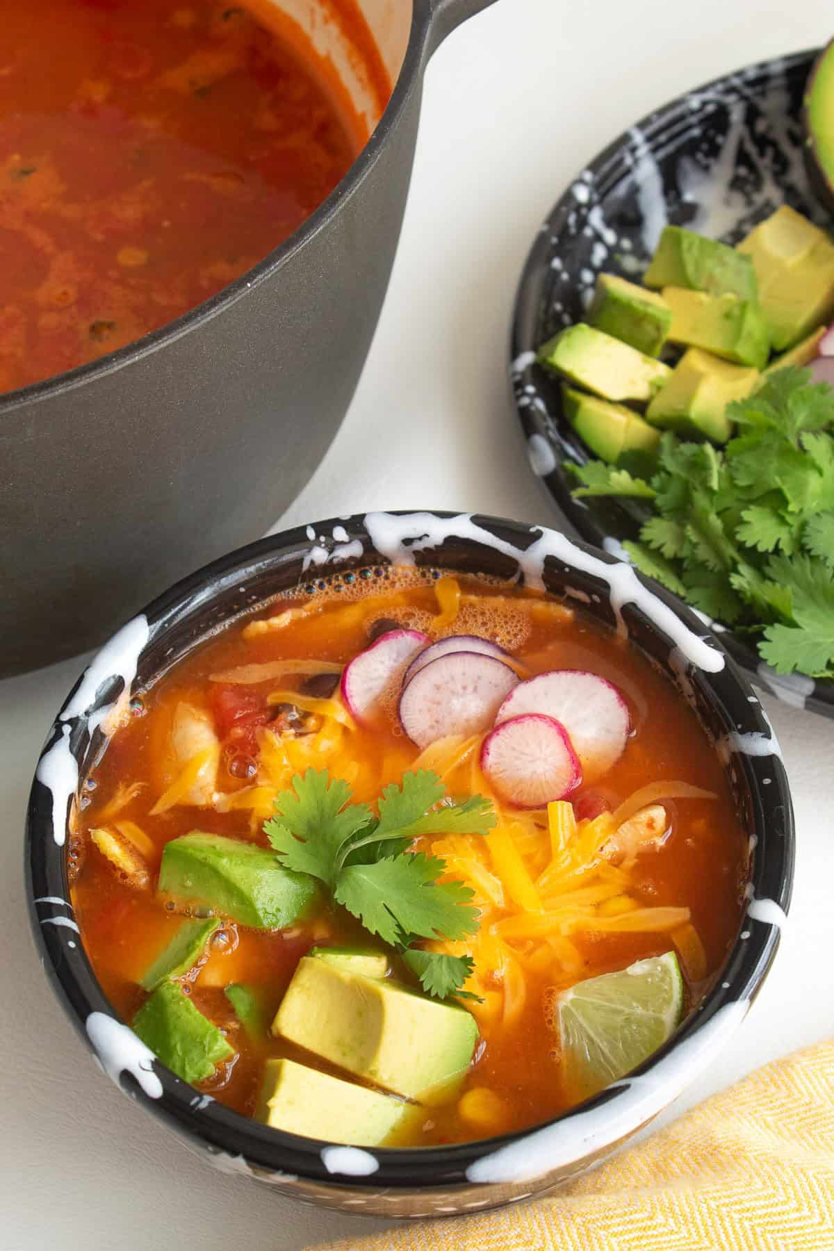 A bowl of garnished brothy soup sits on a white surface alongside the pot of soup and a plate of toppings.