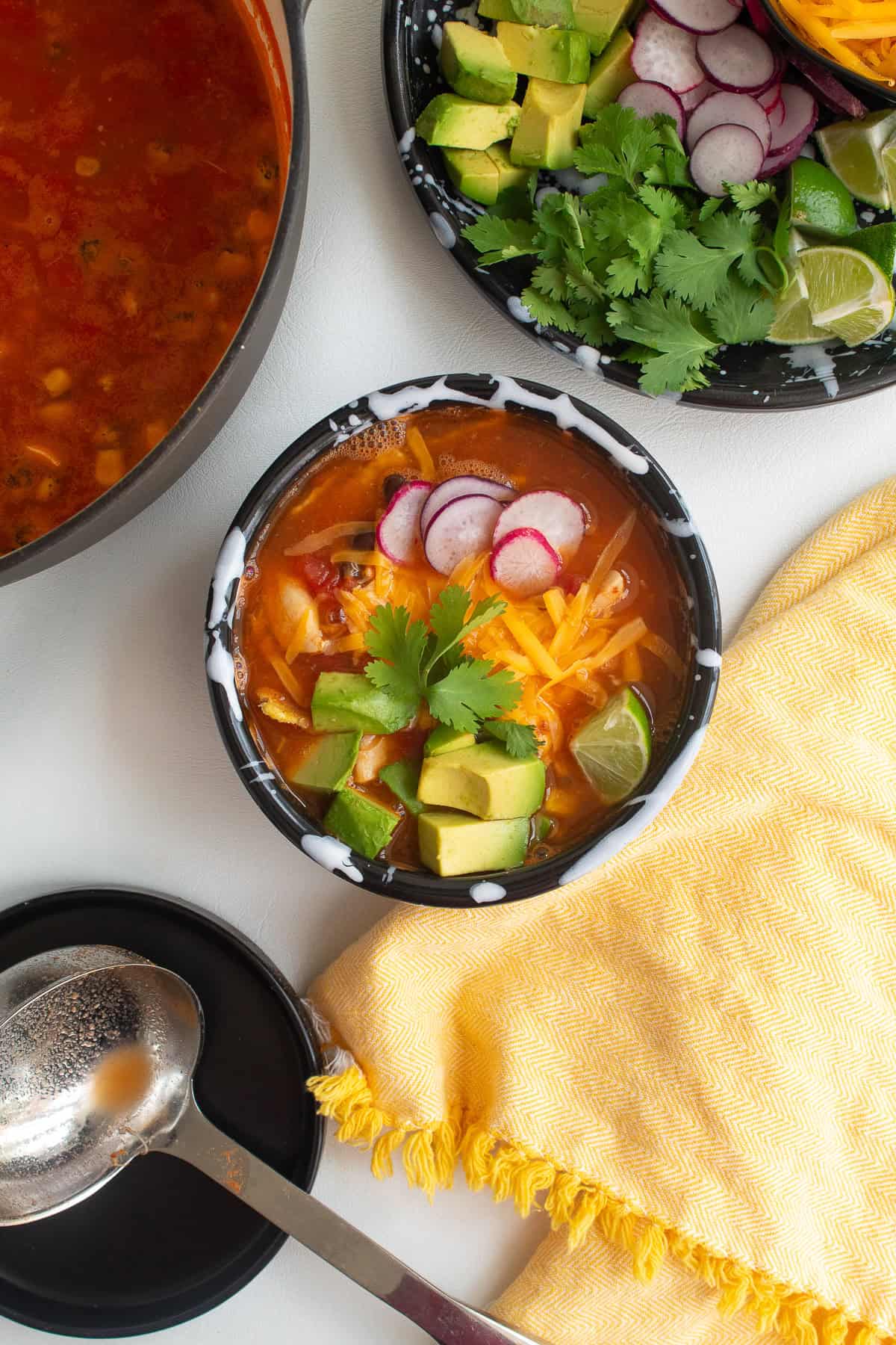 The soup is topped with radish slices, avocado chunks, shredded cheese, cilantro, and a wedge of lime.