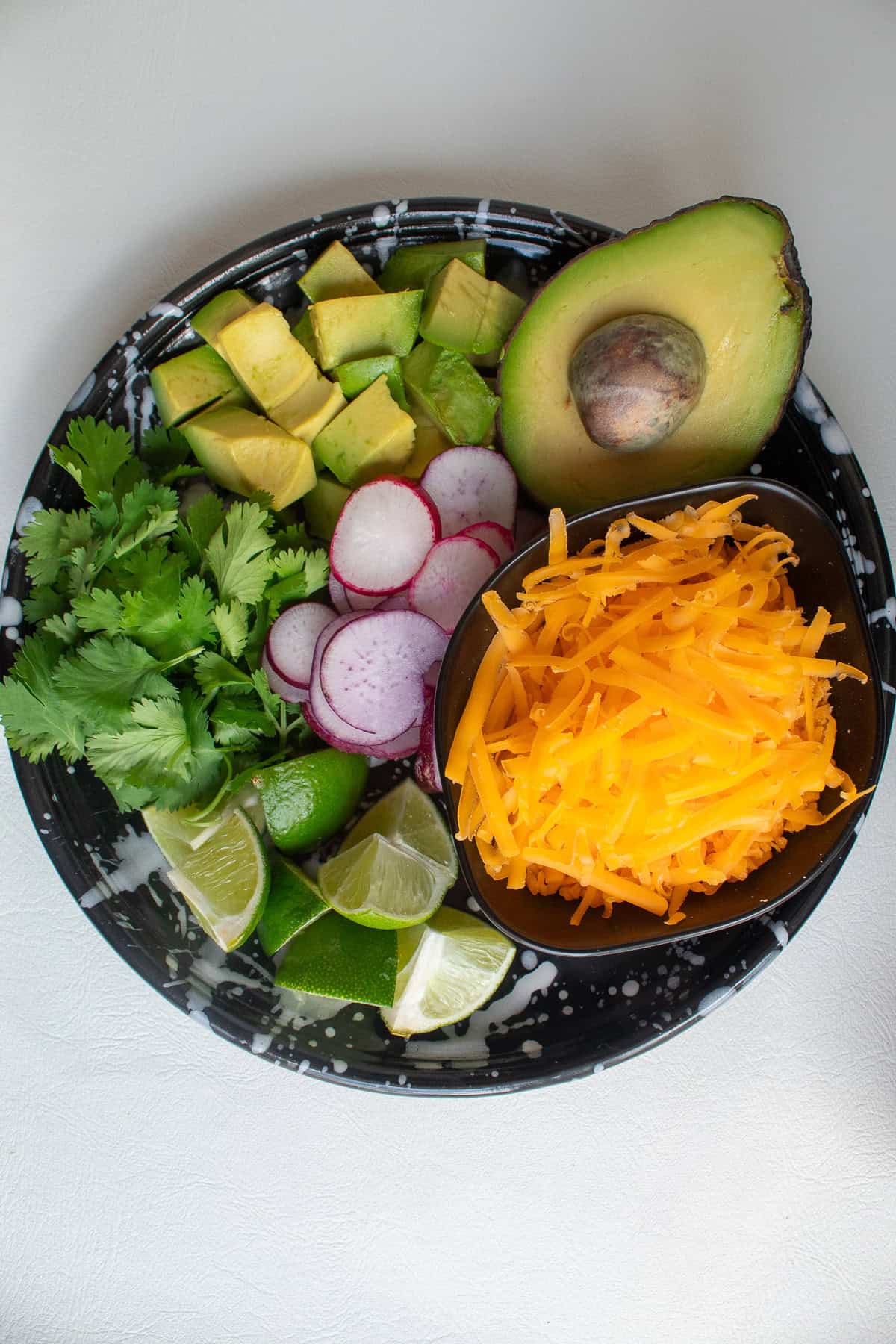 A black plate is piled with toppings for the soup, including avocado, radish, cheese, lime, and cilantro.