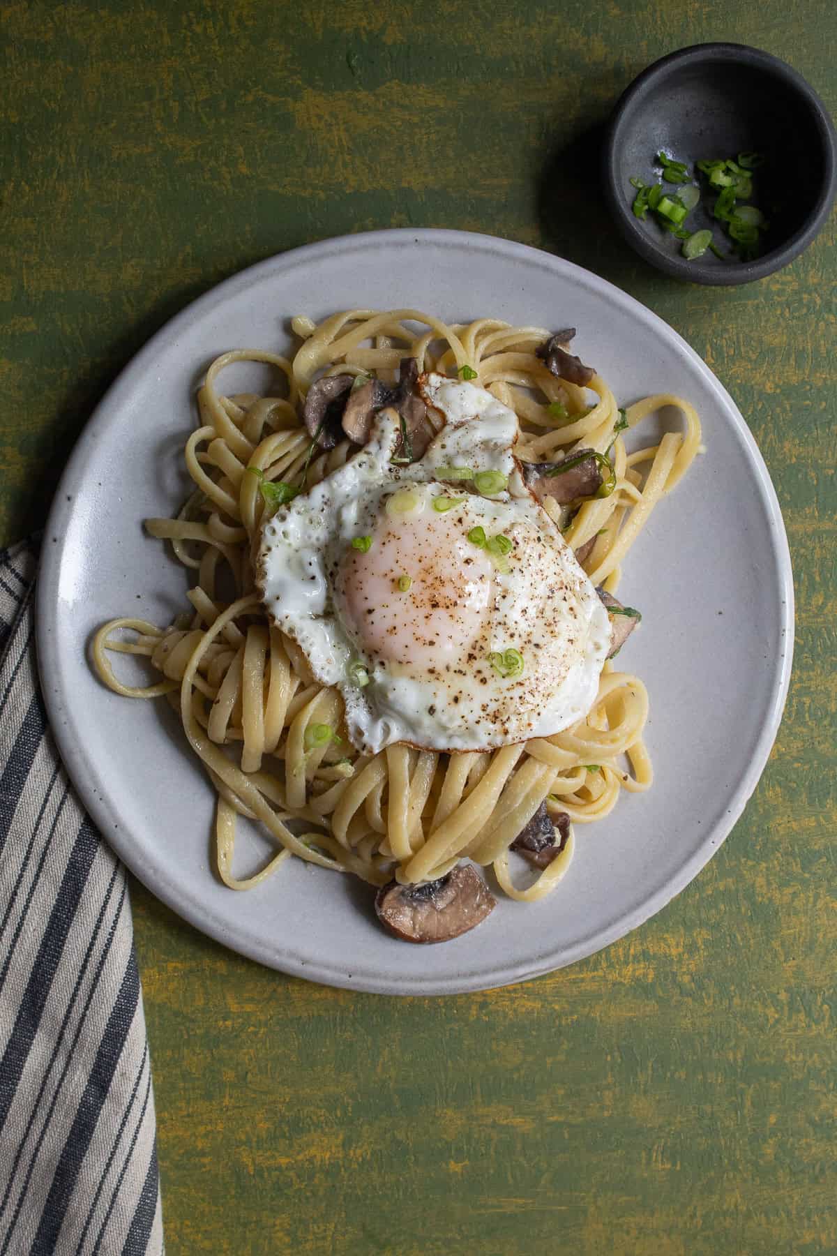 A plate full of pasta is topped with a fried egg that has been generously seasoned with pepper.