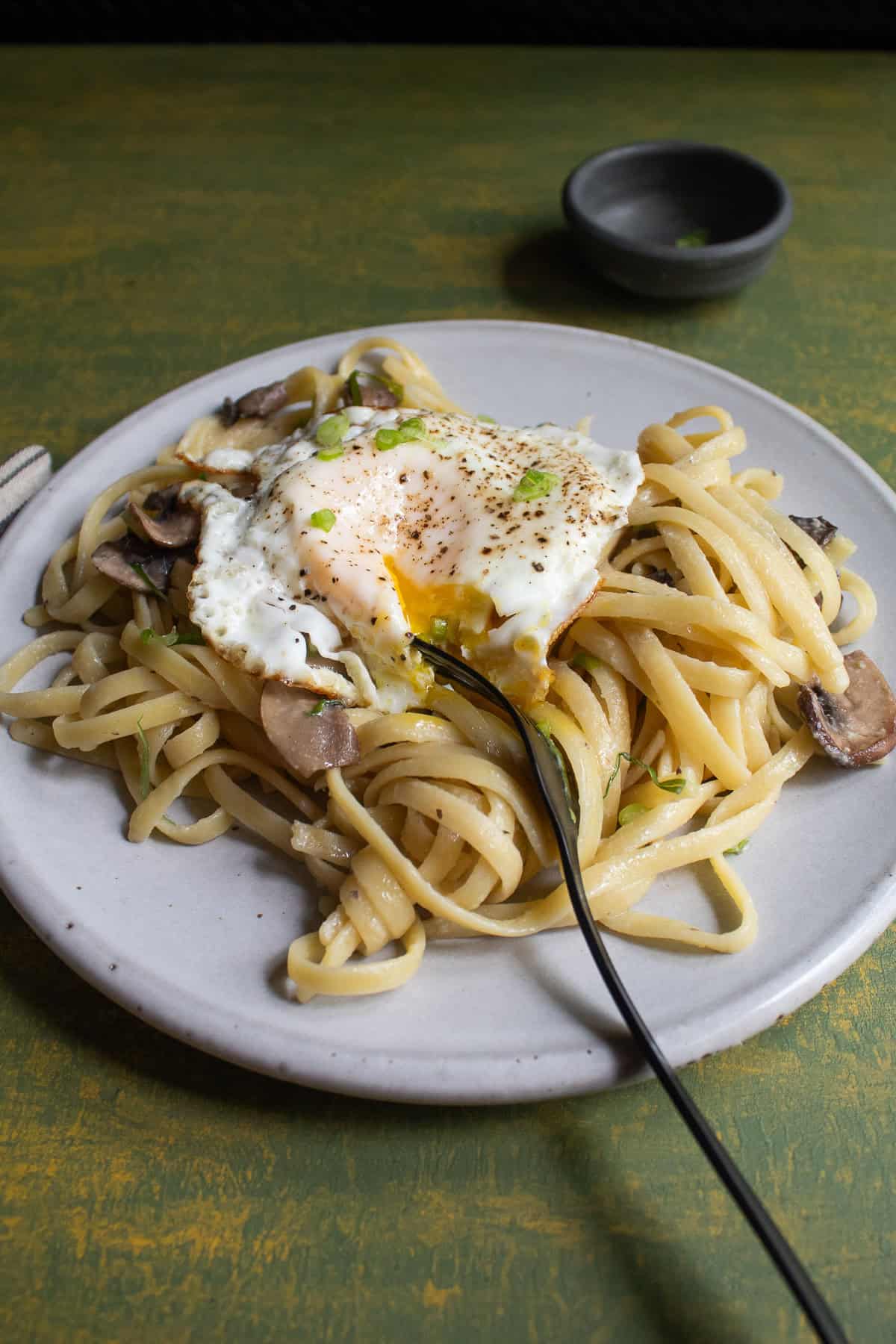 A fried egg is cut open on top of a tangle of fettucine pasta with mushrooms.
