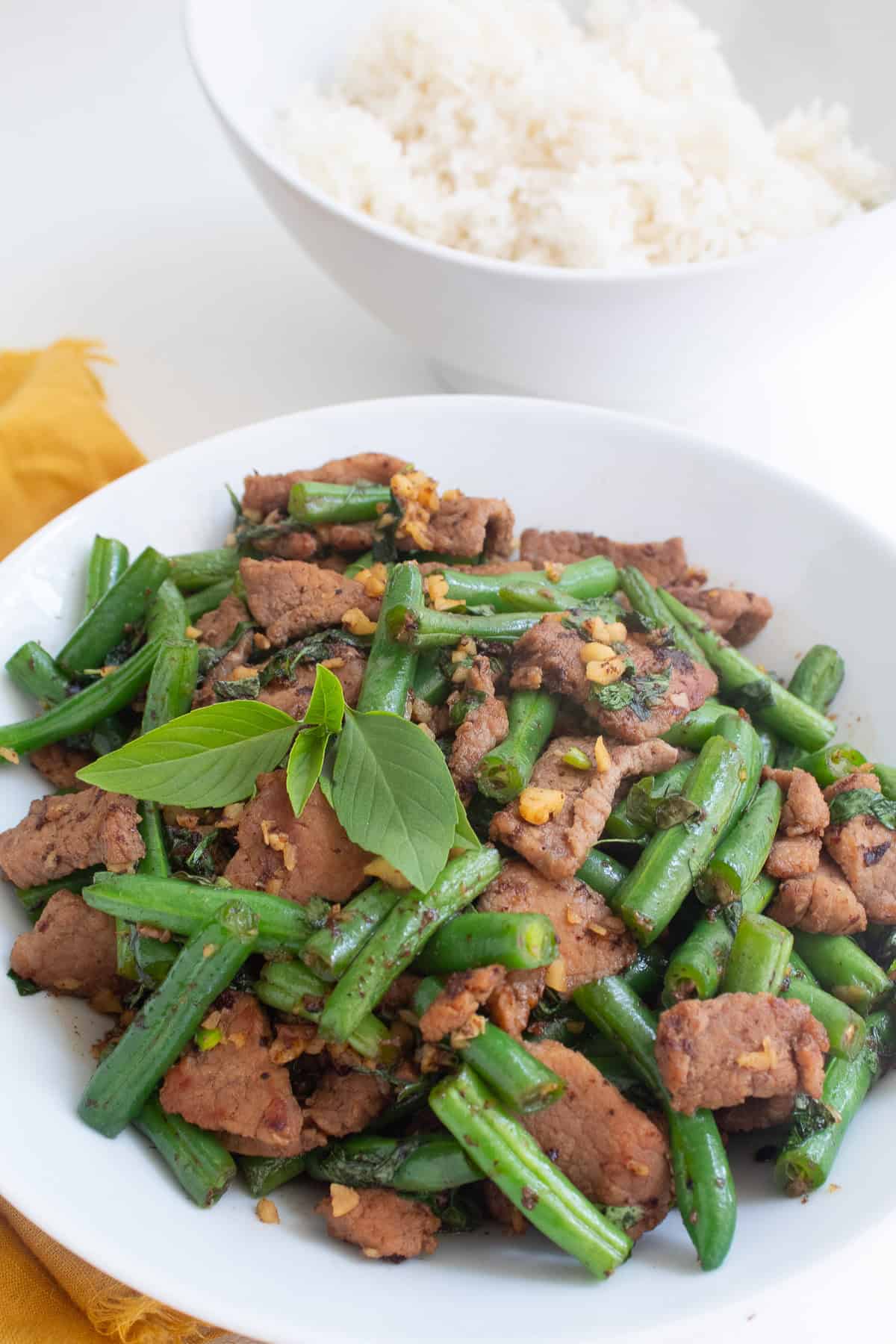 A white bowl of stir fried beef, green beans, garlic, and Thai basil leaves.