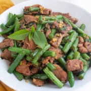 A white bowl of stir fried beef, green beans, garlic, and Thai basil leaves.