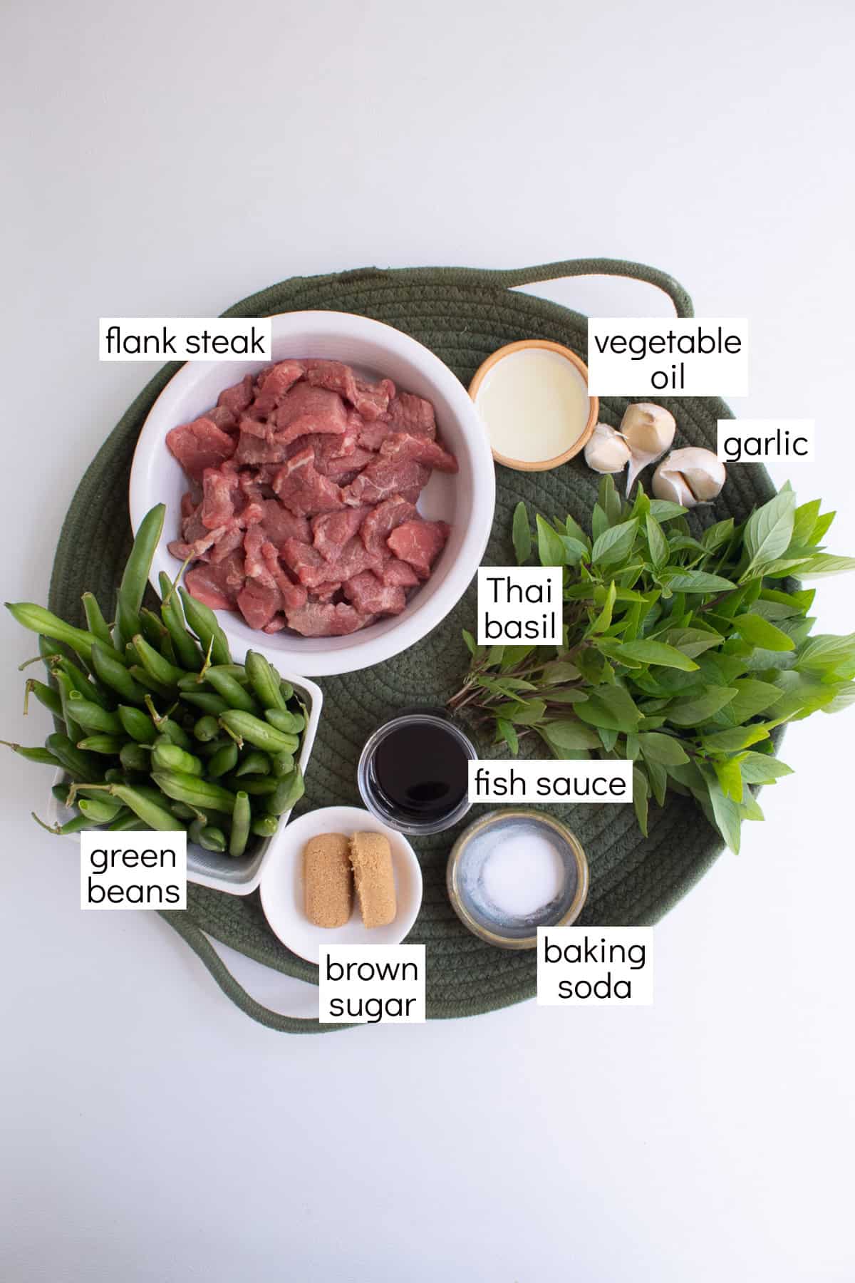 Ingredients for the recipe are displayed on a green platter and are labeled with white text boxes including flank steak, vegetable oil, garlic, Thai basil, fish sauce, green beans, brown sugar, and baking soda.