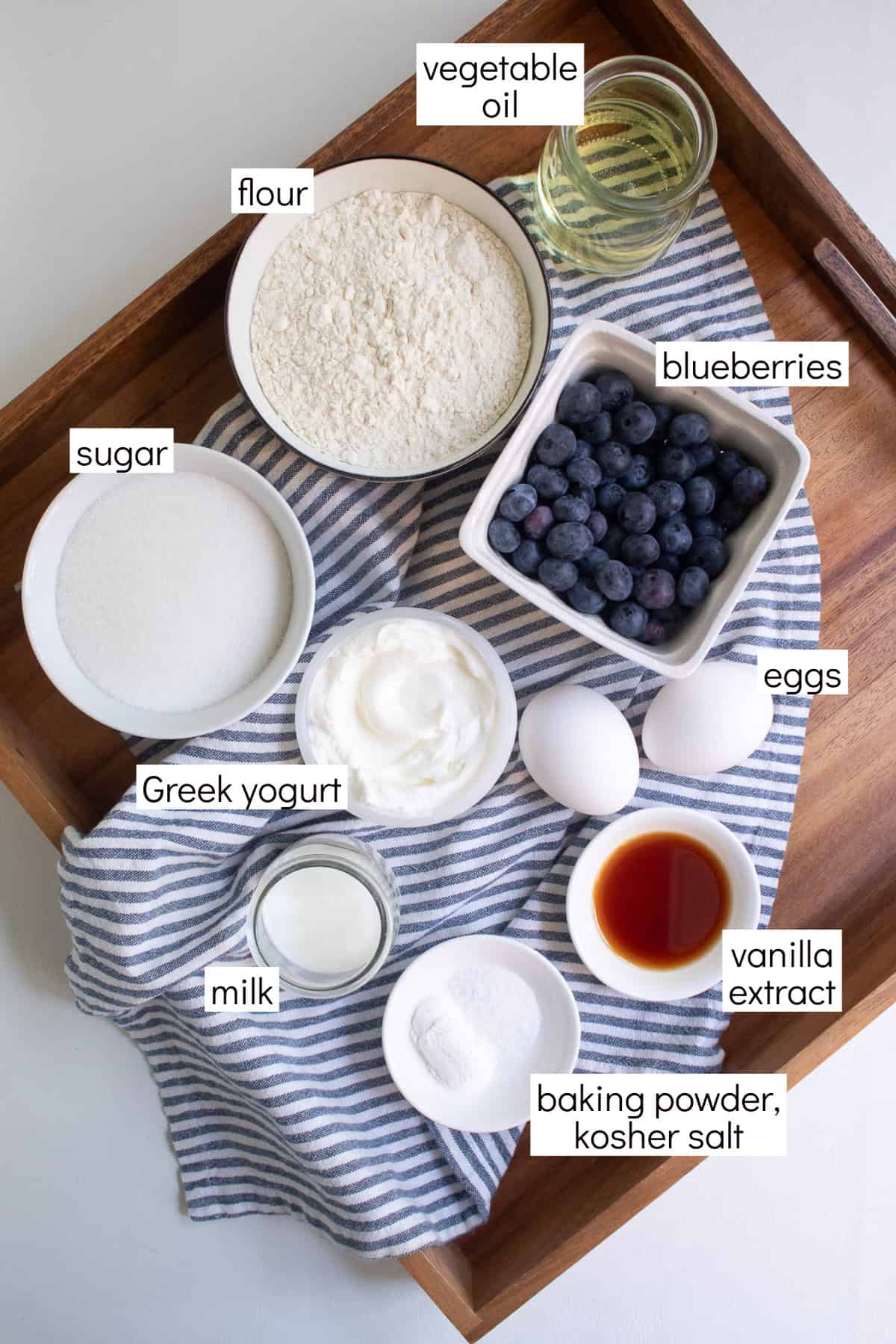 Ingredients for the blueberry coffee cake are displayed in small bowls over a blue and white striped towel and are labeled with white text boxes including vegetable oil, flour, blueberries, sugar, Greek yogurt, eggs, milk, vanilla extract, and baking powder, salt.