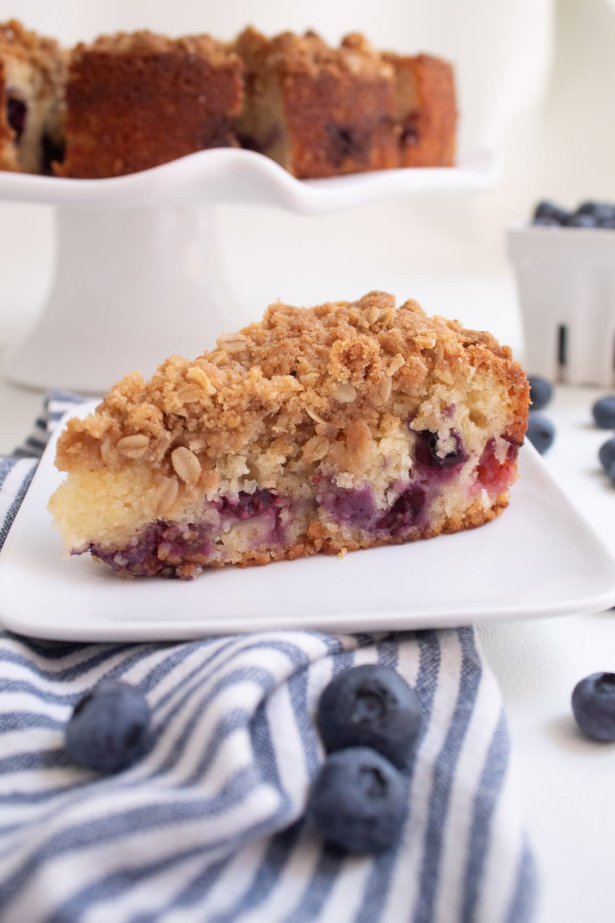 A detail image of a slice of coffee cake shows burst purple blueberries under a layer of golden oat crumble.
