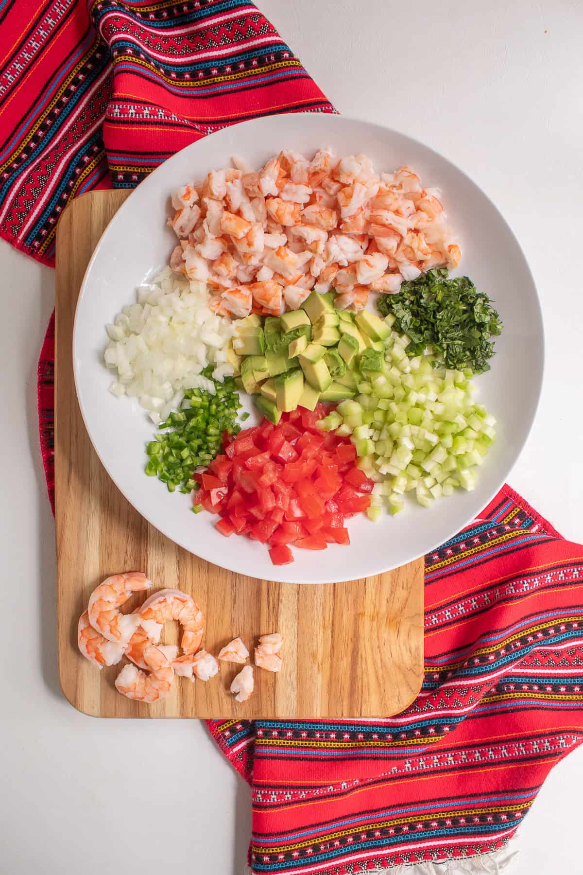 The chopped and diced vegetables sit in a white bowl next to the shrimp on a cutting board.