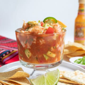 A deep goblet contains a red mixture with visible shrimp, tomato, and avocado chunks and lime and jalapeño garnishes.