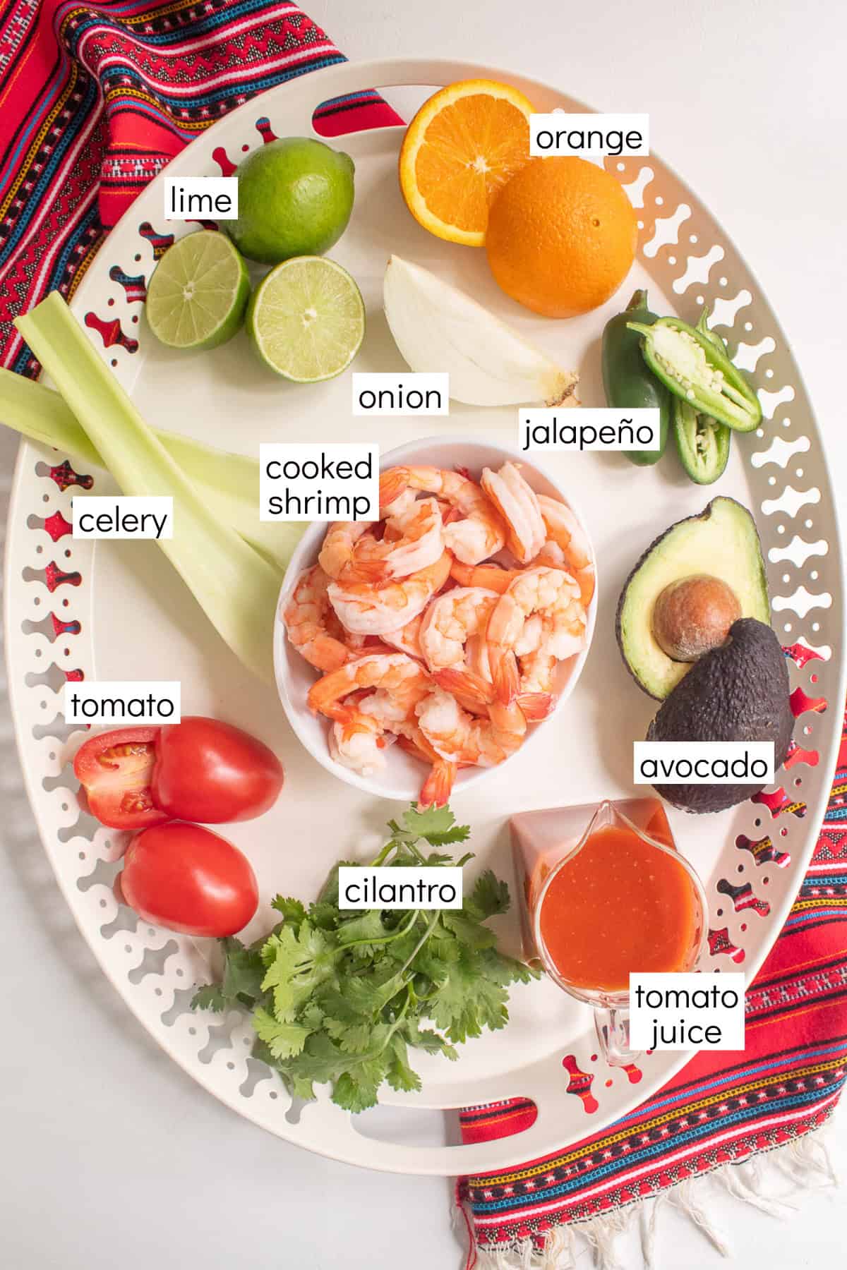 Ingredients for the recipe are displayed and labeled with white text boxes including the words lime, orange, onion, jalapeño, celery, cooked shrimp, tomato, avocado, cilantro, and tomato juice.
