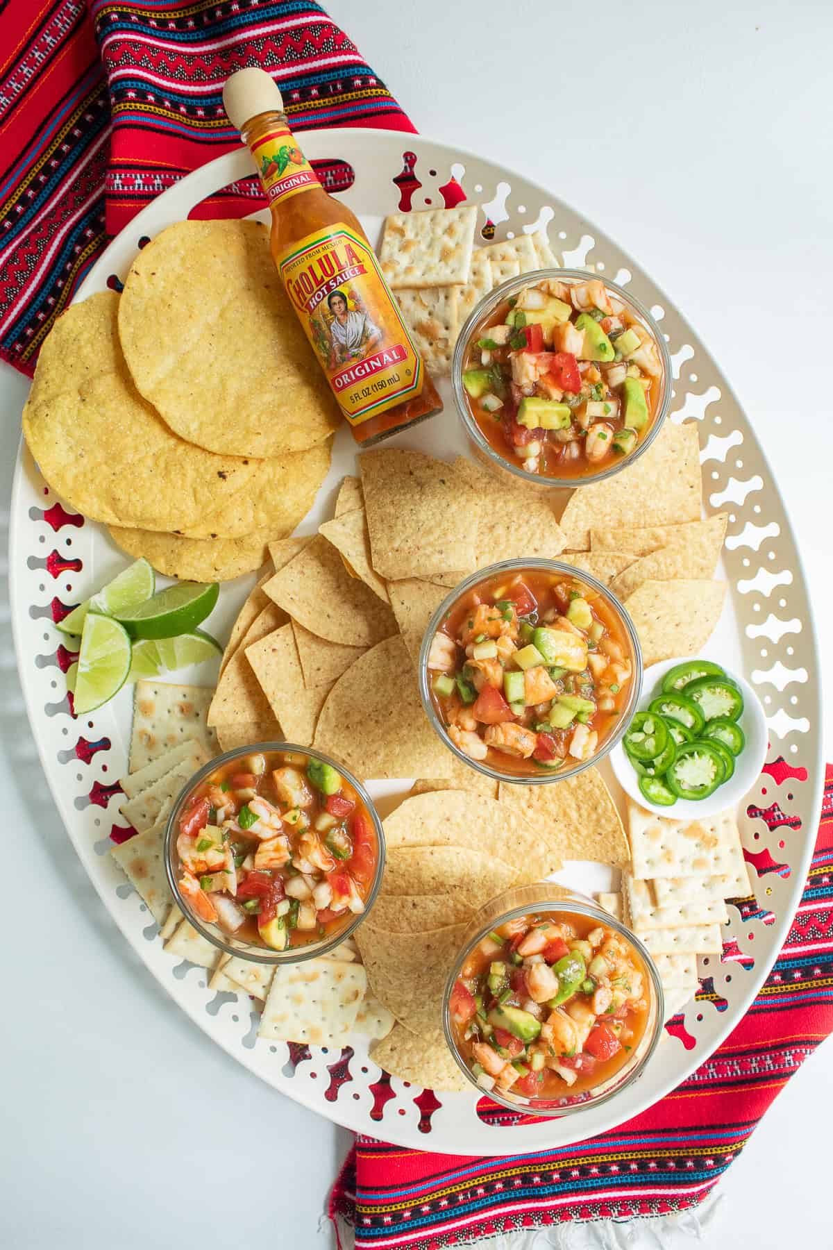 A party platter holds tostadas, tortilla chips, saltines, lime wedges, hot sauce, jalapeño rings, and bowls of shrimp cocktail.
