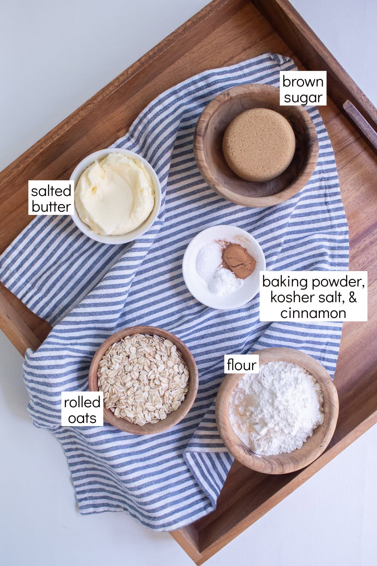 Ingredients for the blueberry coffee cake are displayed in small bowls over a blue and white striped towel and are labeled with white text boxes including brown sugar, salted butter, baking powder, kosher salt, & cinnamon, rolled oats, and flour.