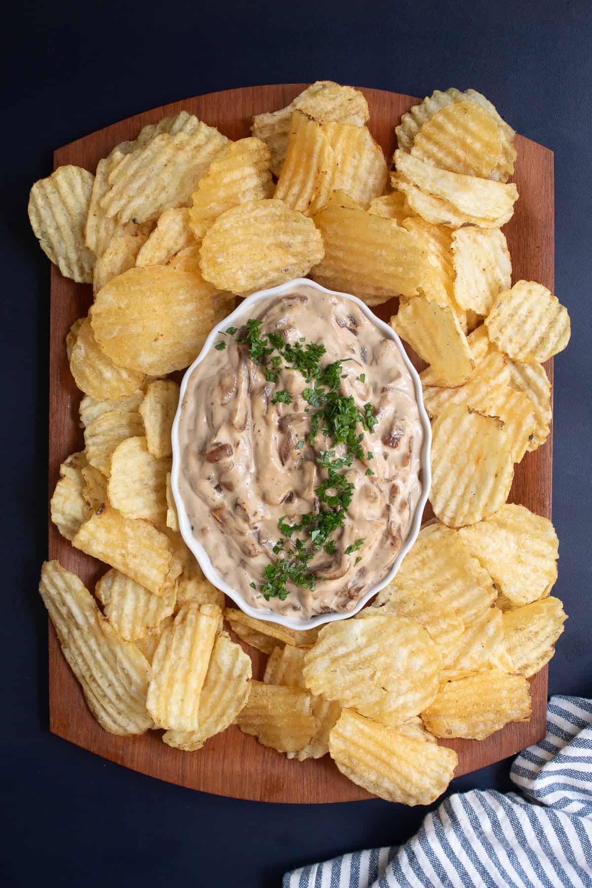 A brown wooden tray has a white bowl of onion dip surrounded by ridged potato chips.