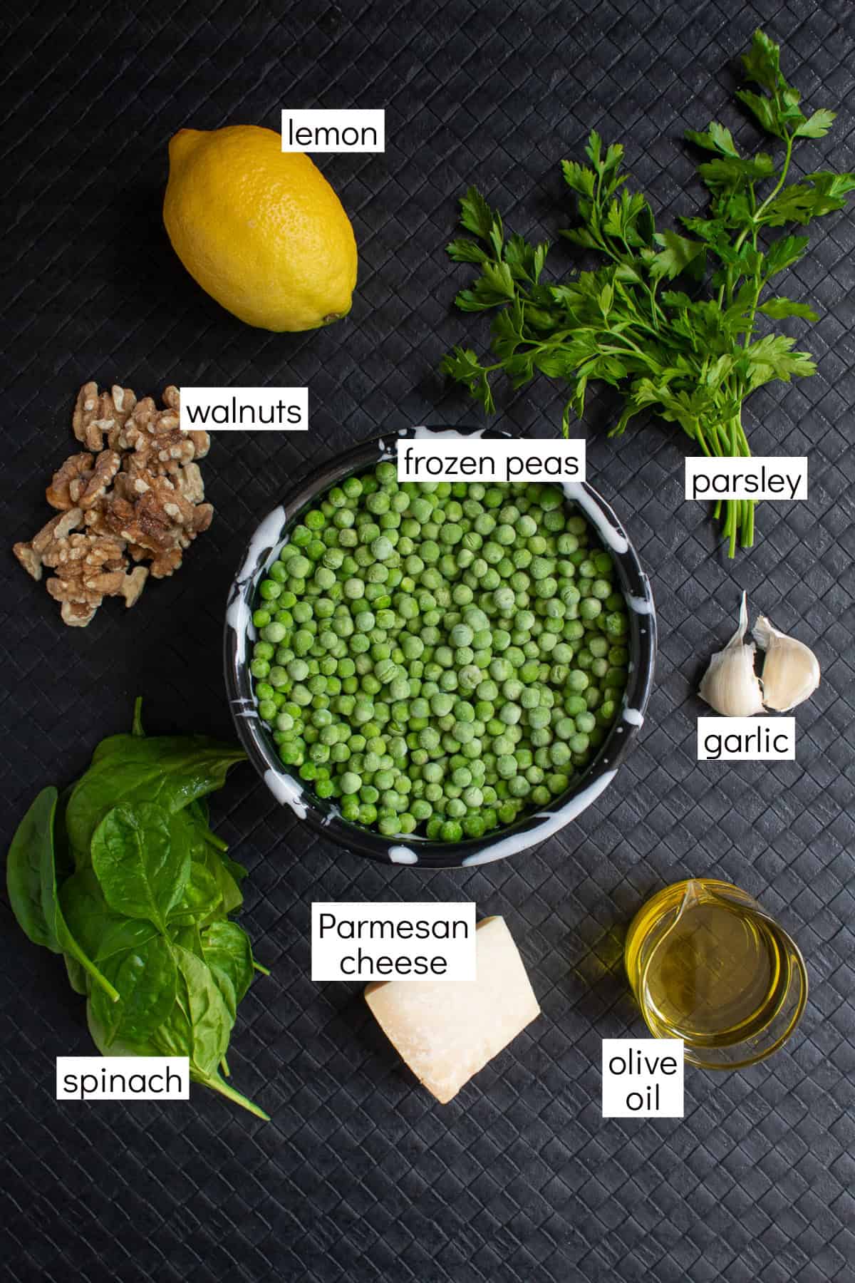 Ingredients for the recipe are displayed on a black surface and are labeled with white text boxes including lemon, walnuts, frozen peas, parsley, garlic, spinach, Parmesan cheese, and olive oil.