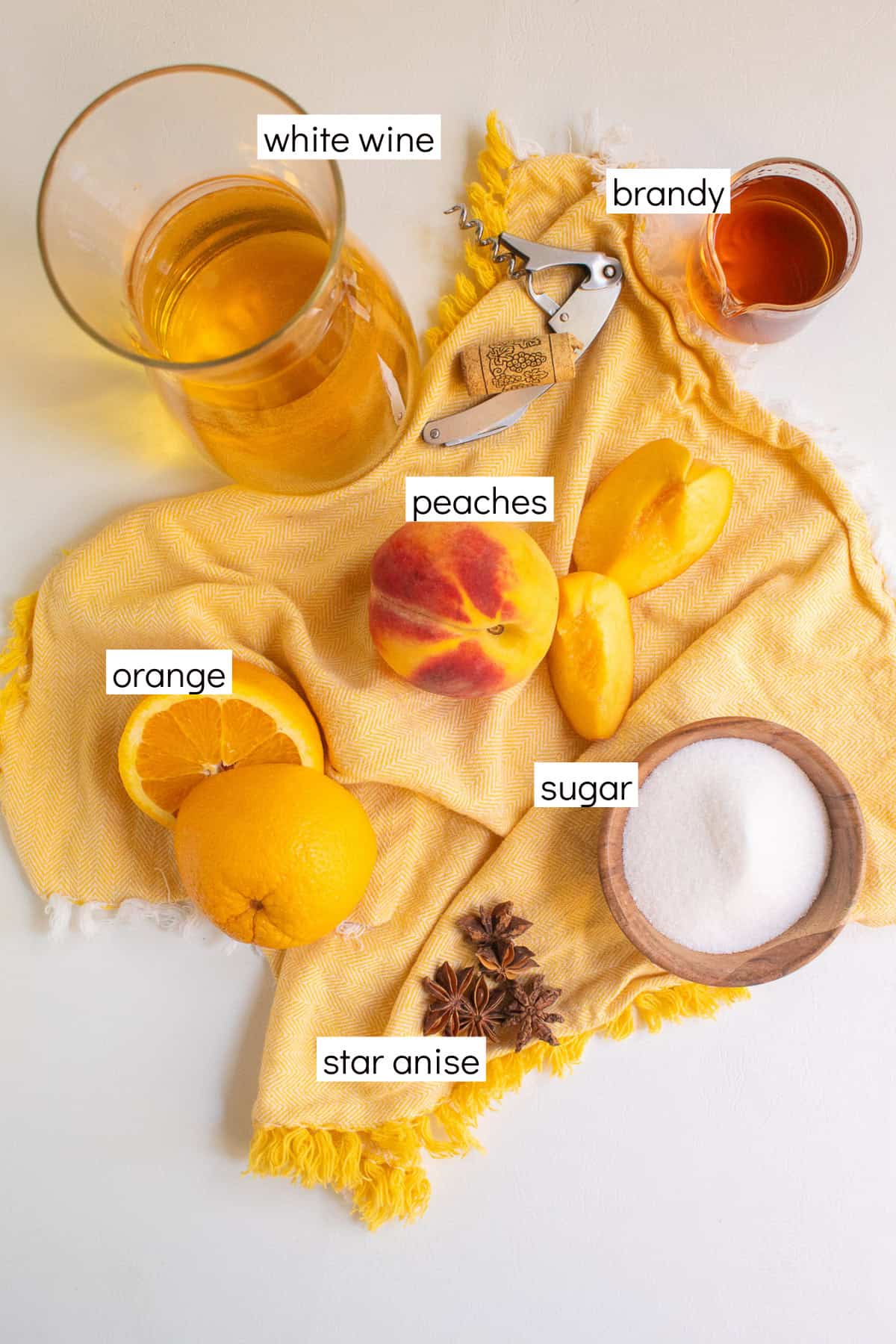 Ingredients for the sangria are displayed on a yellow surface with white text box labels including white wine, brandy, peaches, orange, sugar, and star anise.