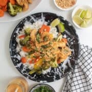 A black and white plate is piled with rice, chicken, veggies, peanut sauce, cilantro, lime wedges, and crushed peanuts.