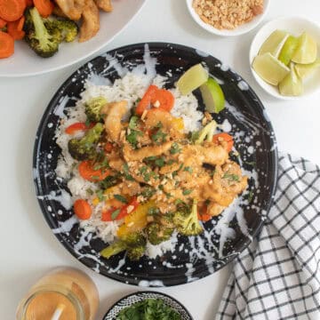 A black and white plate is piled with rice, chicken, veggies, peanut sauce, cilantro, lime wedges, and crushed peanuts.