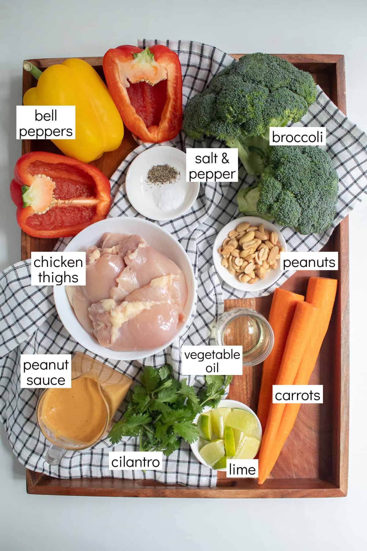 Ingredients for the recipe are displayed on a wood tray and are labeled with white text boxes including bell peppers, salt & pepper, broccoli, chicken thighs, peanuts, peanut sauce, vegetable oil, carrots, cilantro, and lime.