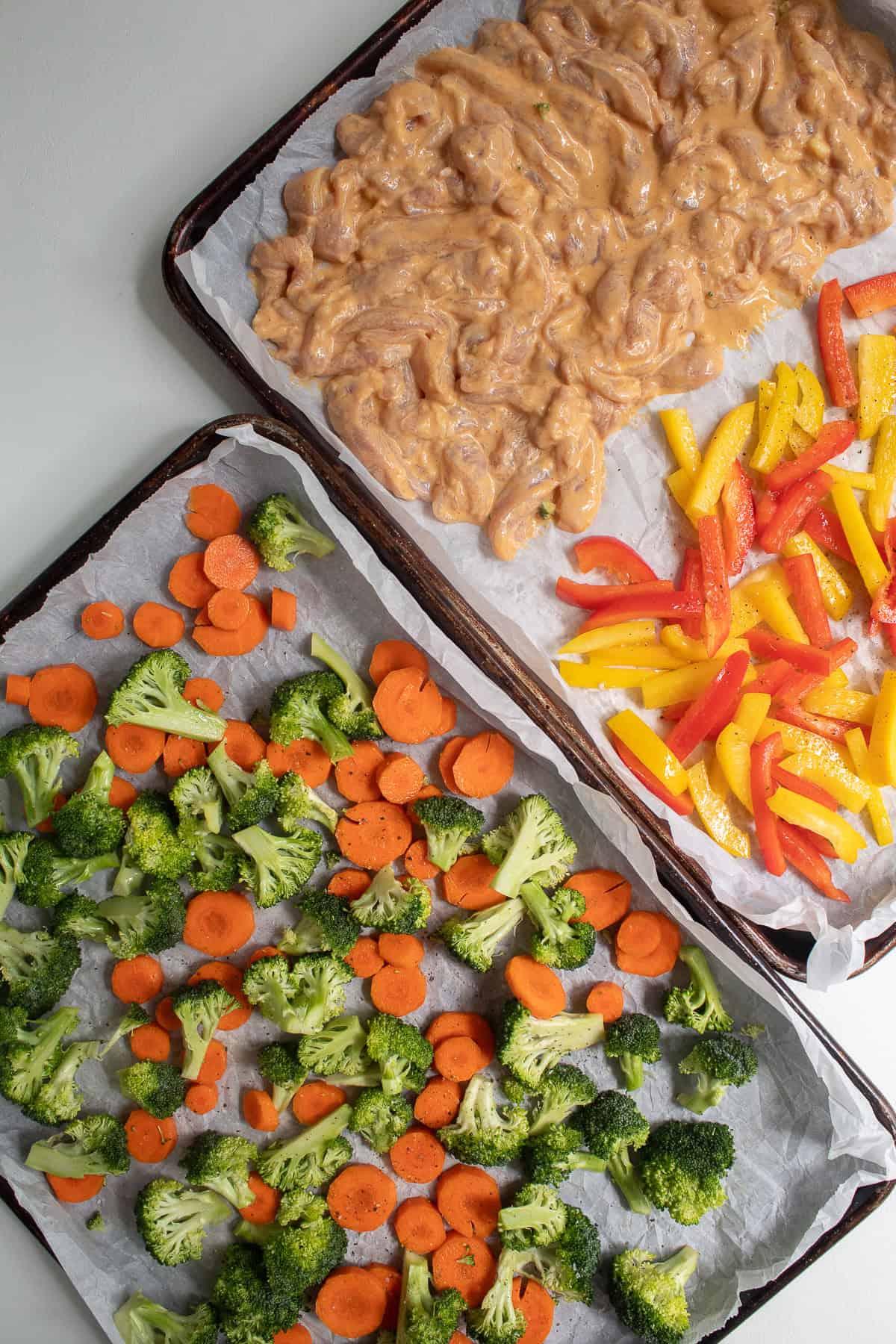 One sheet pan is loaded with broccoli and carrots, while a second sheet pan has marinated chicken and red and yellow peppers.