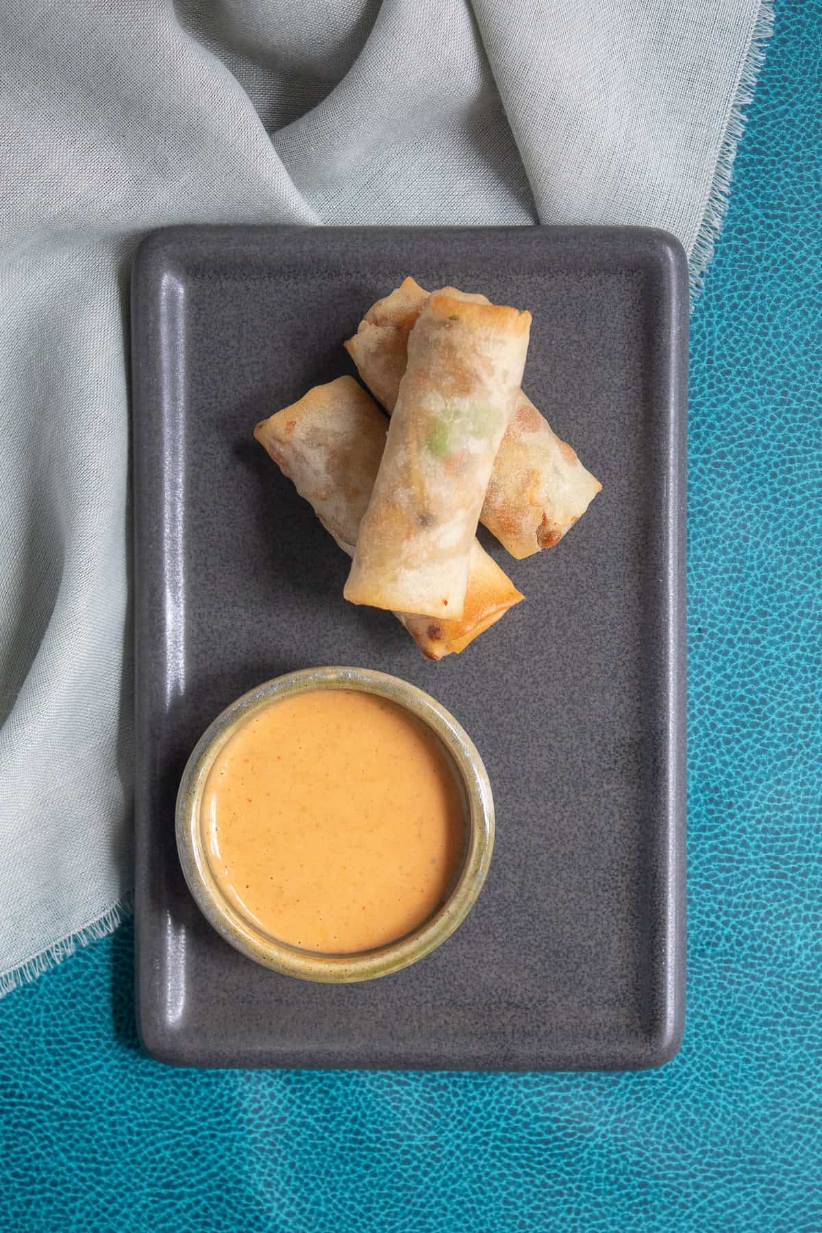 Crispy spring rolls are on a gray plate next to a small bowl of Thai peanut sauce for dipping.