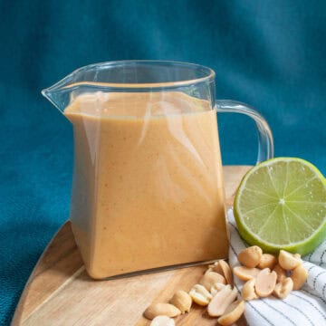 A pitcher of peanut sauce sits next to roasted peanuts and a lime slice on a wood board.