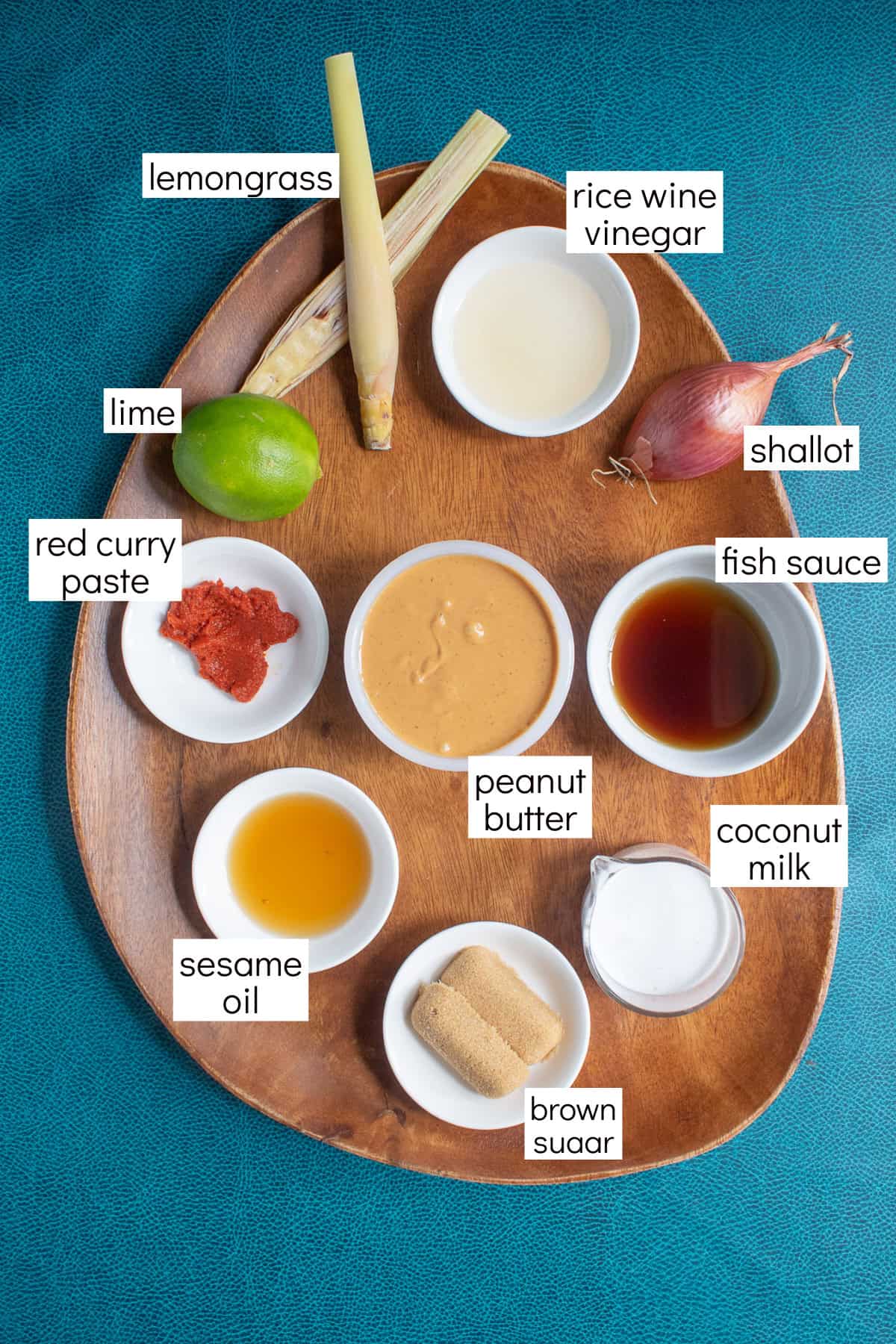 Ingredients for the recipe are displayed on a wooden platter and are labeled with white text boxes including lemongrass, rice wine vinegar, shallot, fish sauce, coconut milk, peanut butter, brown sugar, sesame oil, red curry paste, and lime.