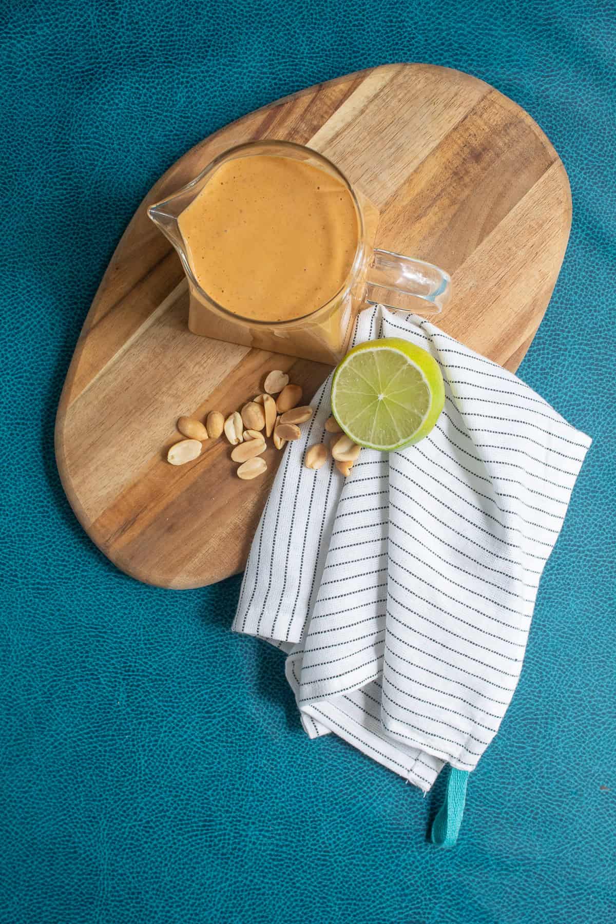 A pitcher of peanut sauce sits next to roasted peanuts and a lime slice on a wood board.