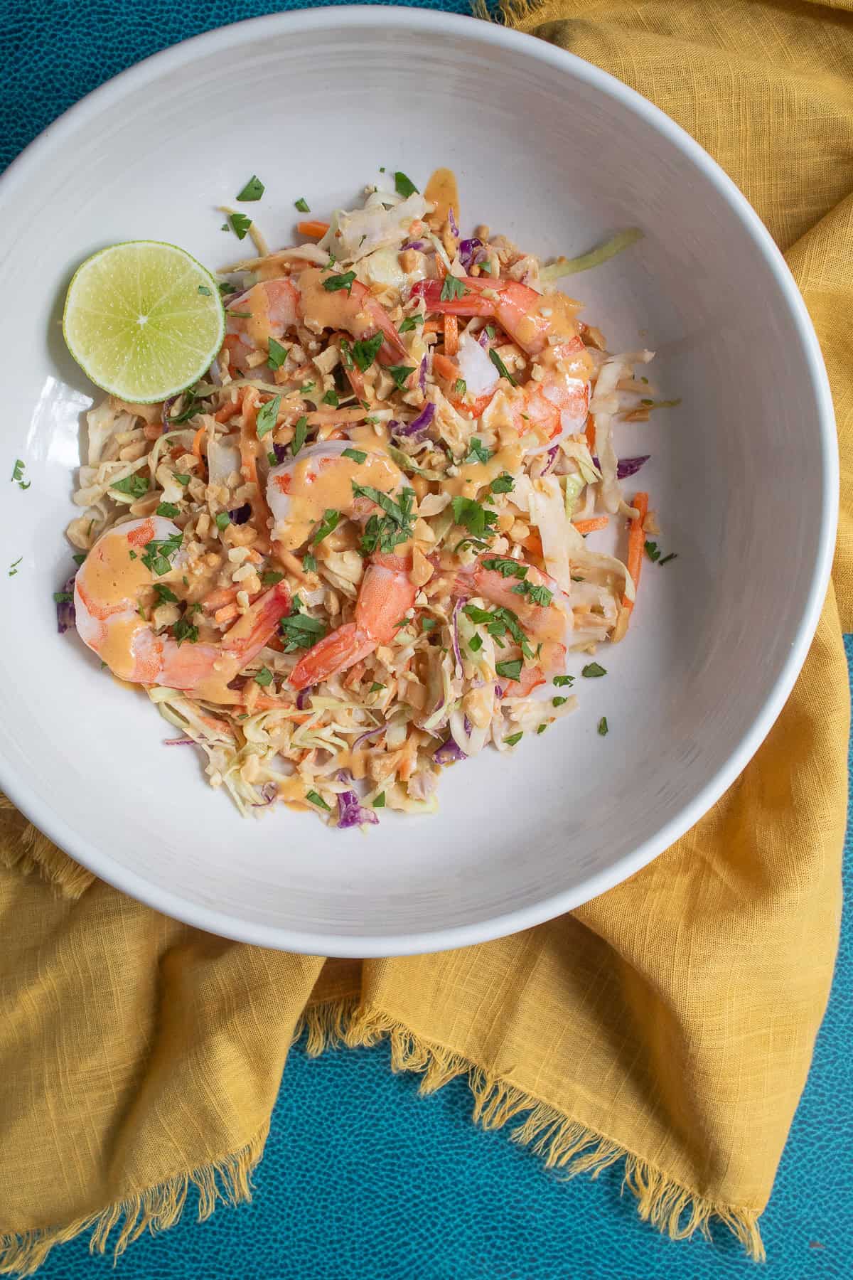 A salad with cabbage, carrots, cilantro, peanuts, and shrimp is dressed with Thai peanut sauce in a white bowl.