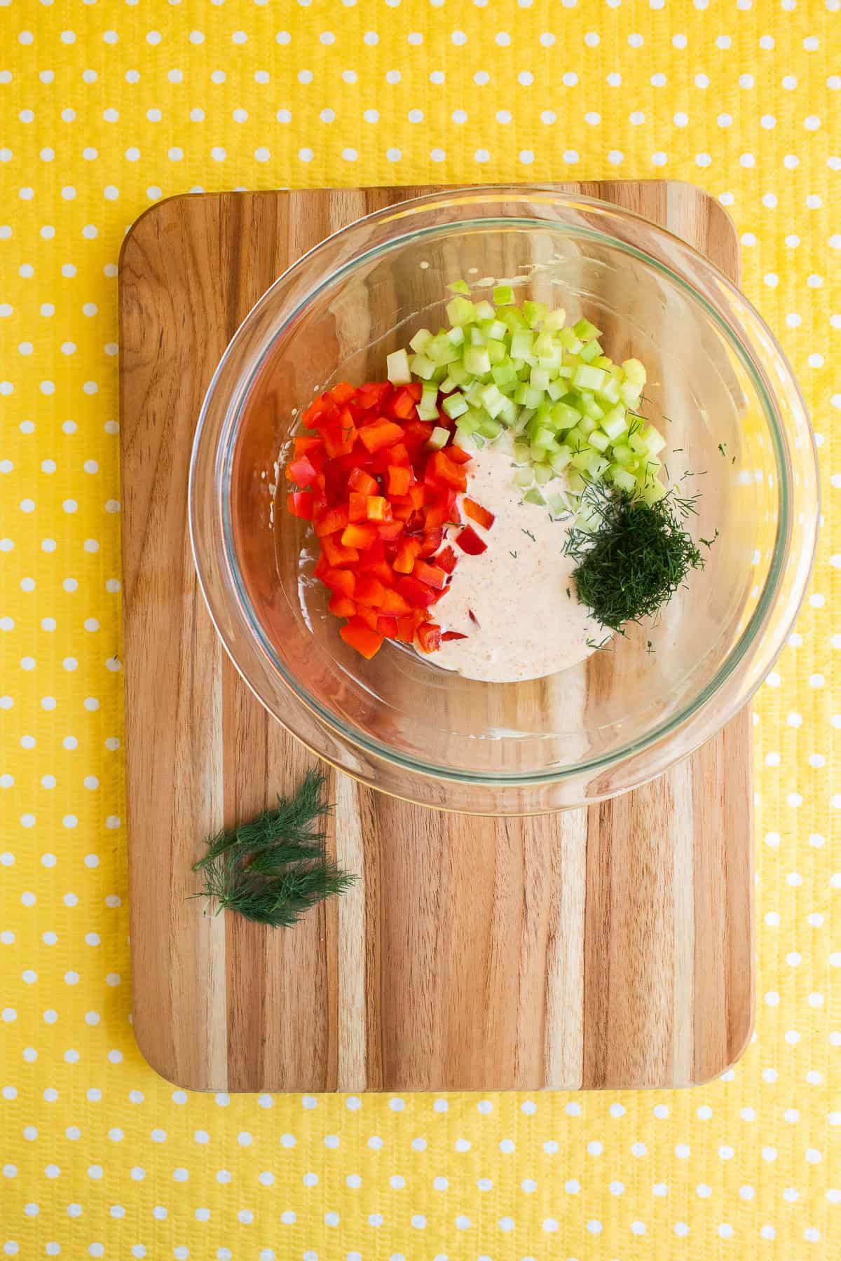A glass bowl contains diced red bell pepper, celery, chopped dill, and a mayonnaise sauce.