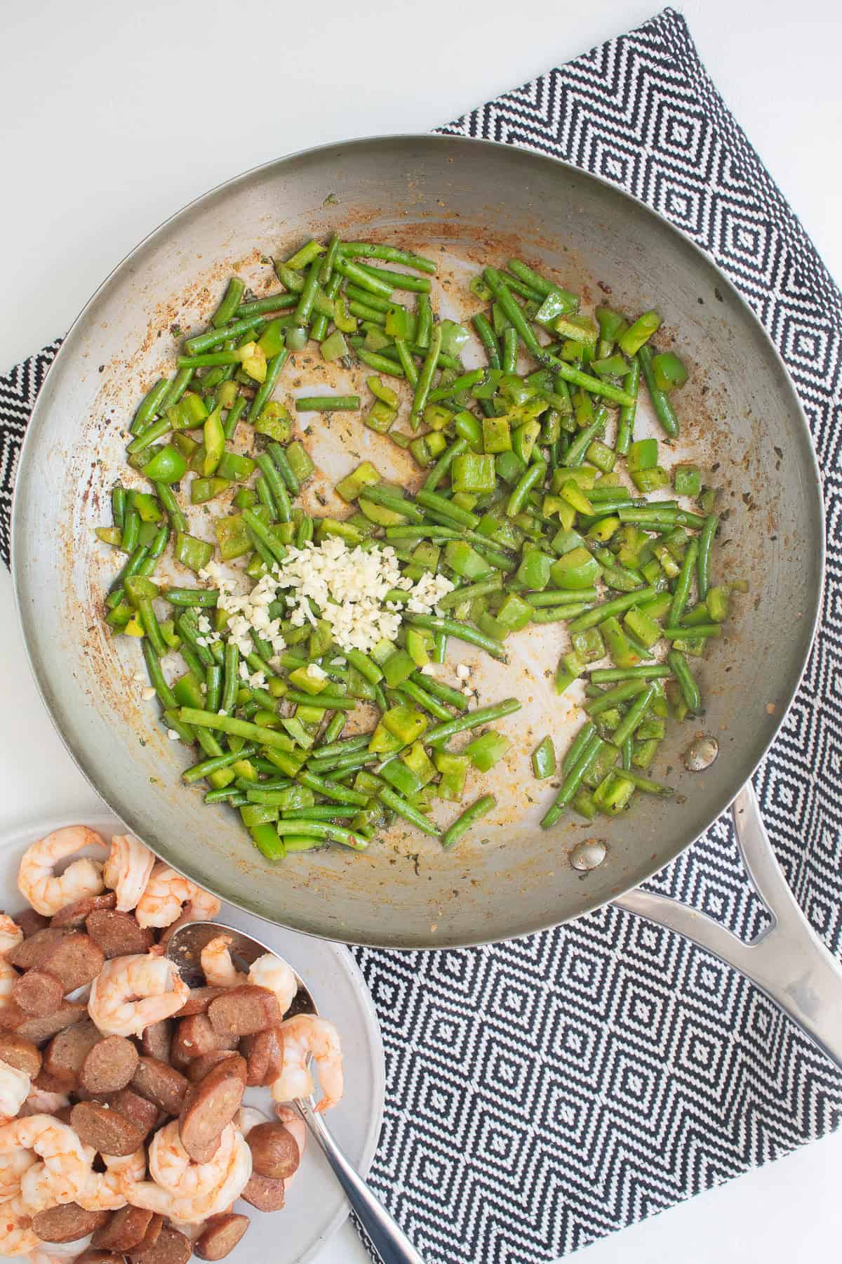 Minced garlic is added to sauteed bell peppers and green beans in a skillet next to a plate of cooked shrimp and sausage.