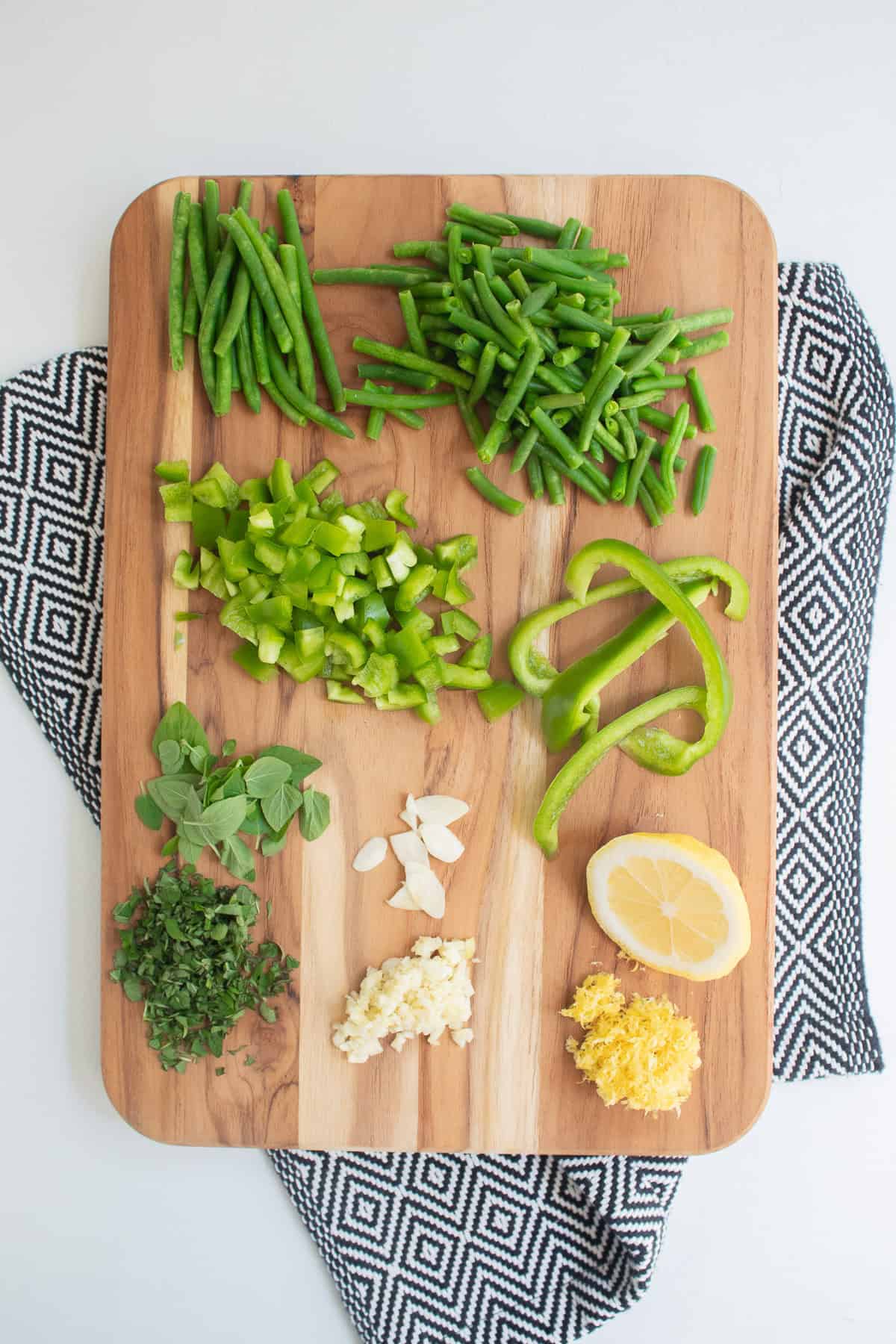 Green beans, bell peppers, oregano, garlic, and lemon are prepared for the dish on a wooden cutting board.